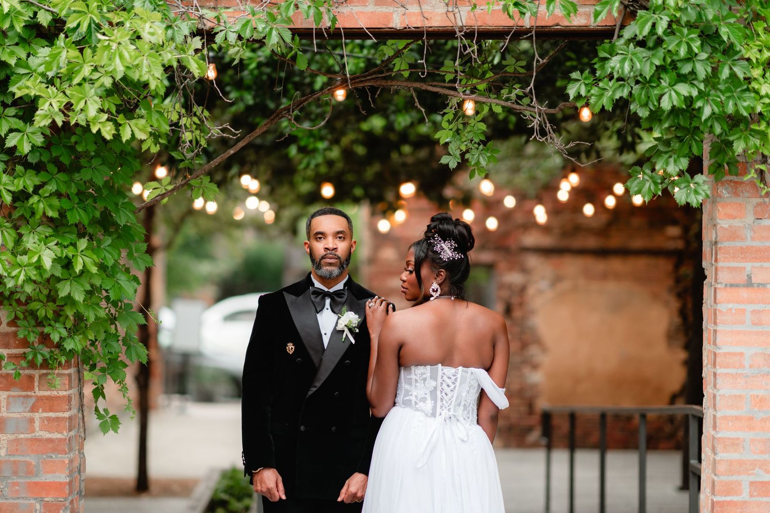 Wedding party members walk along a brick pathway with green foliage and string lights overhead.