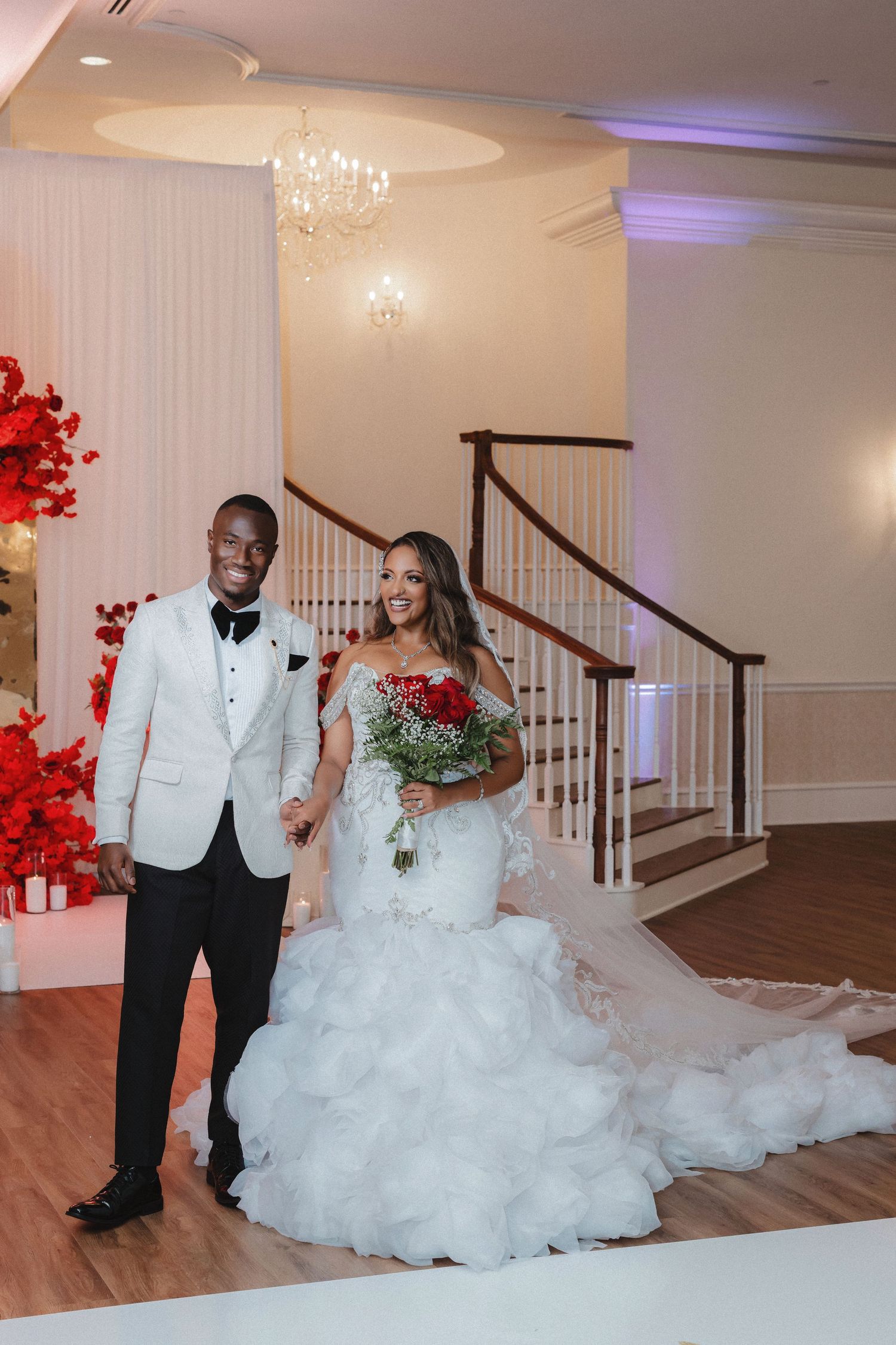 Wedding couple walking together in formal attire with dramatic red floral decorations in the background.