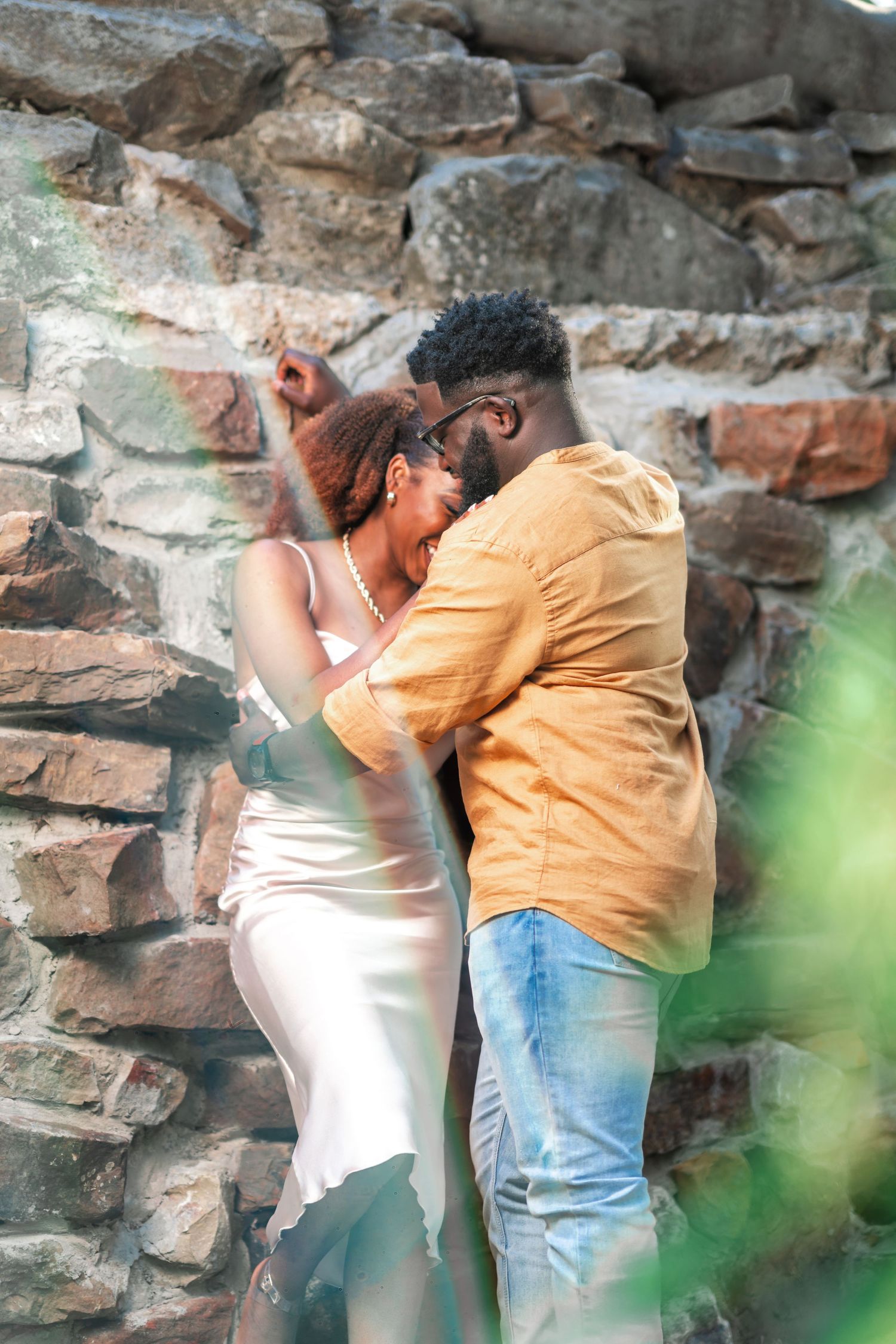 A couple embraces near a stone wall during an outdoor photoshoot.