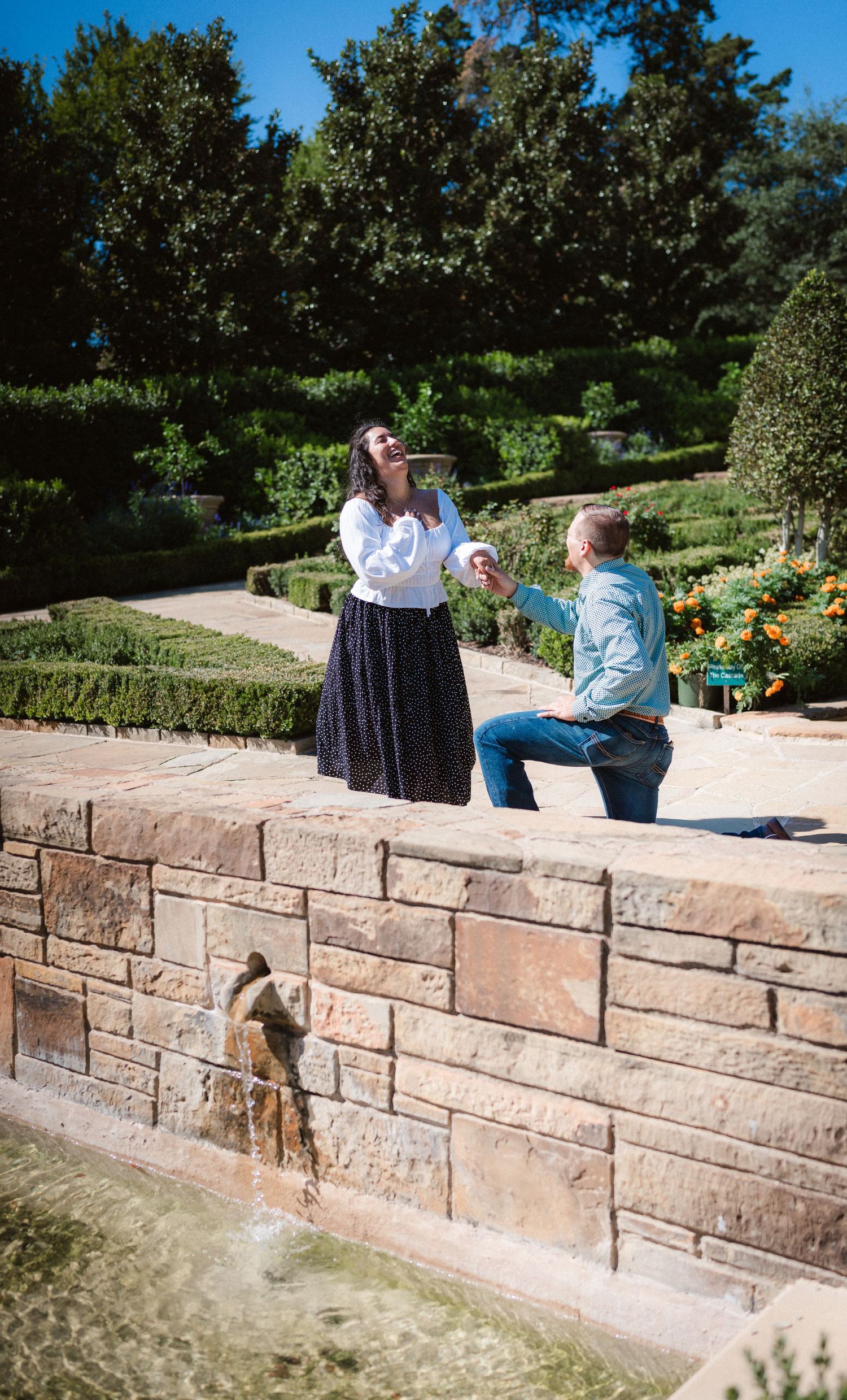 A joyful reaction to a marriage proposal in a manicured garden with a stone fountain wall.
