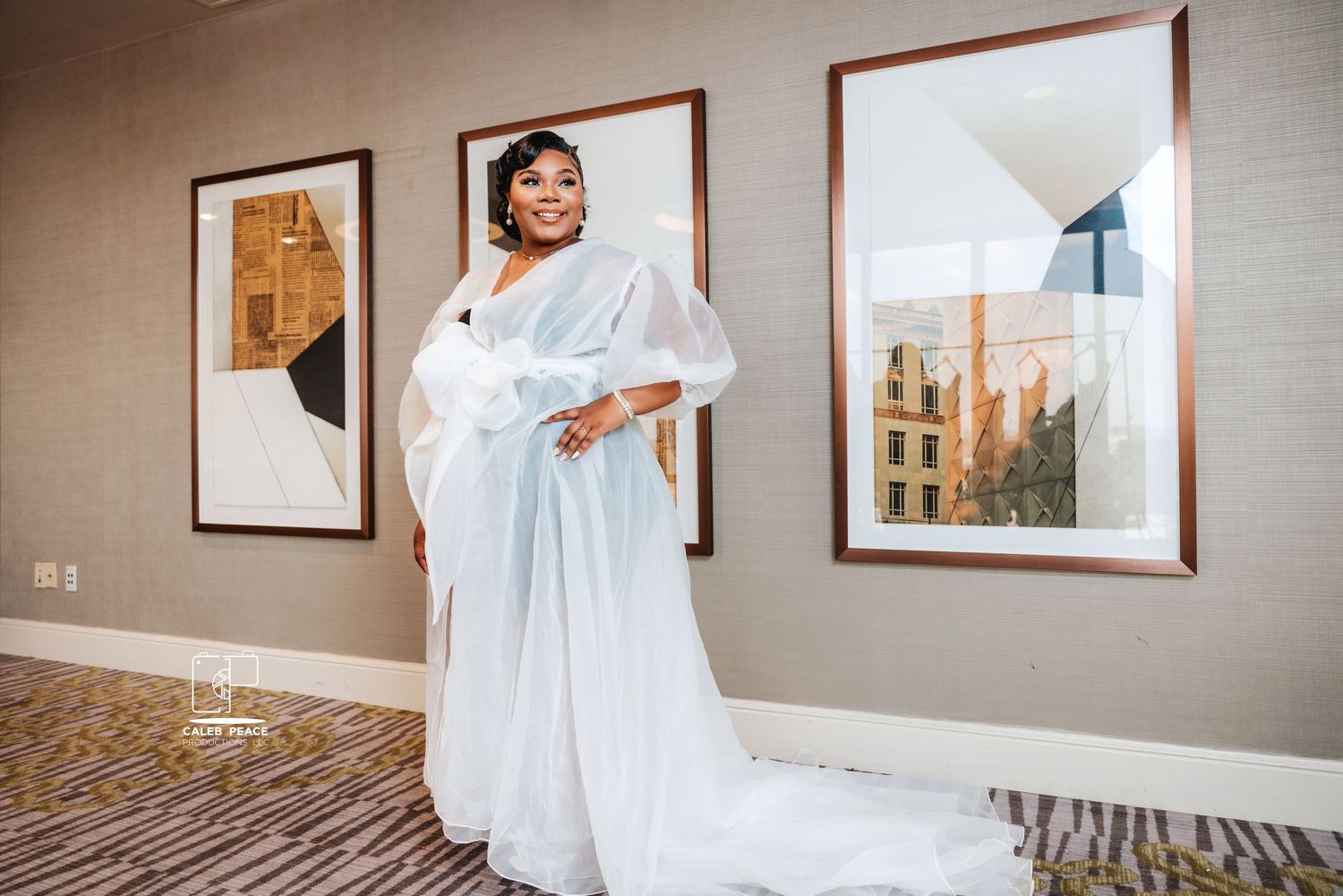 A person wearing an elegant white flowing bridal gown stands in front of framed artwork in a modern interior setting.