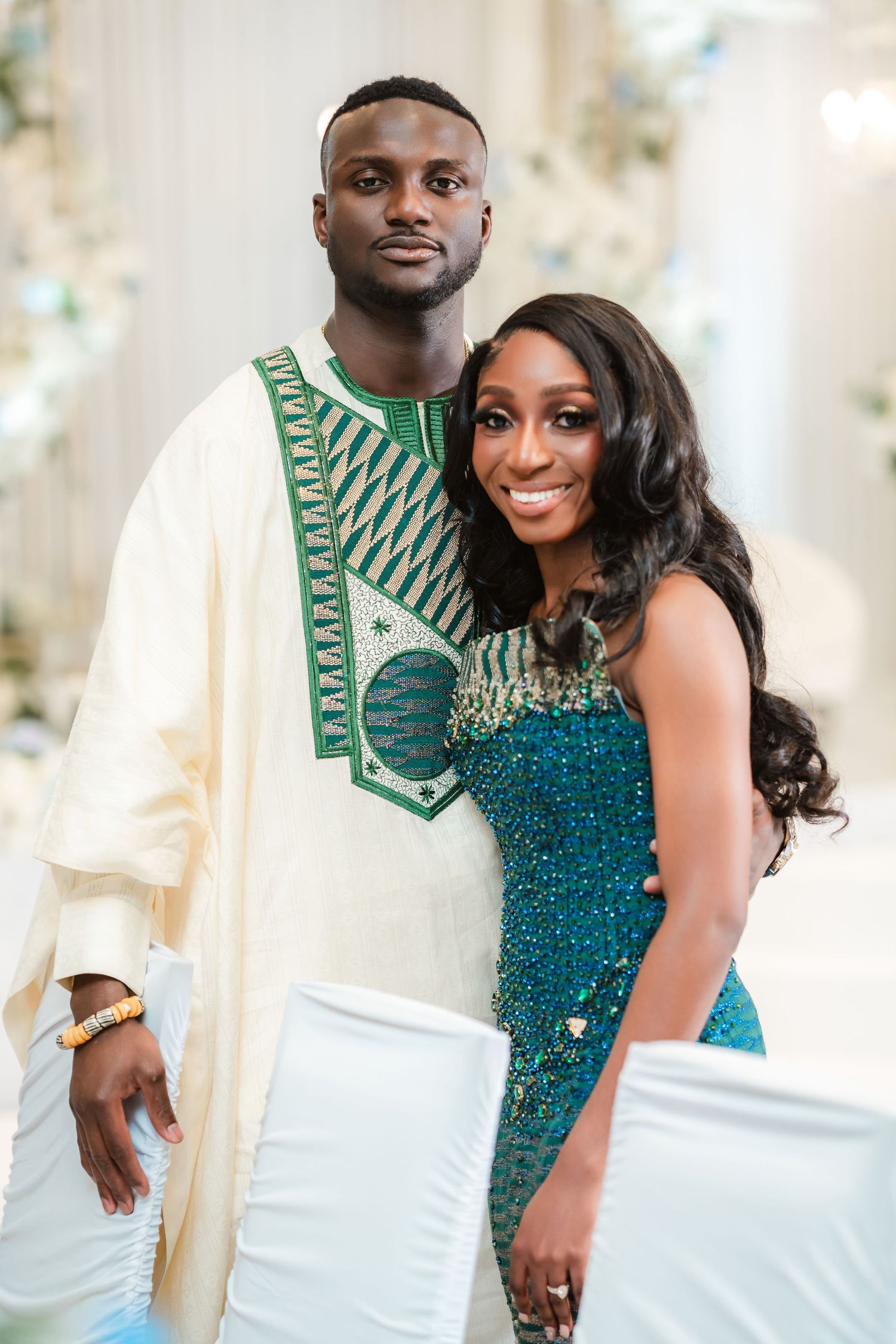 A couple poses together in formal attire featuring a white agbada and sparkly teal gown.
