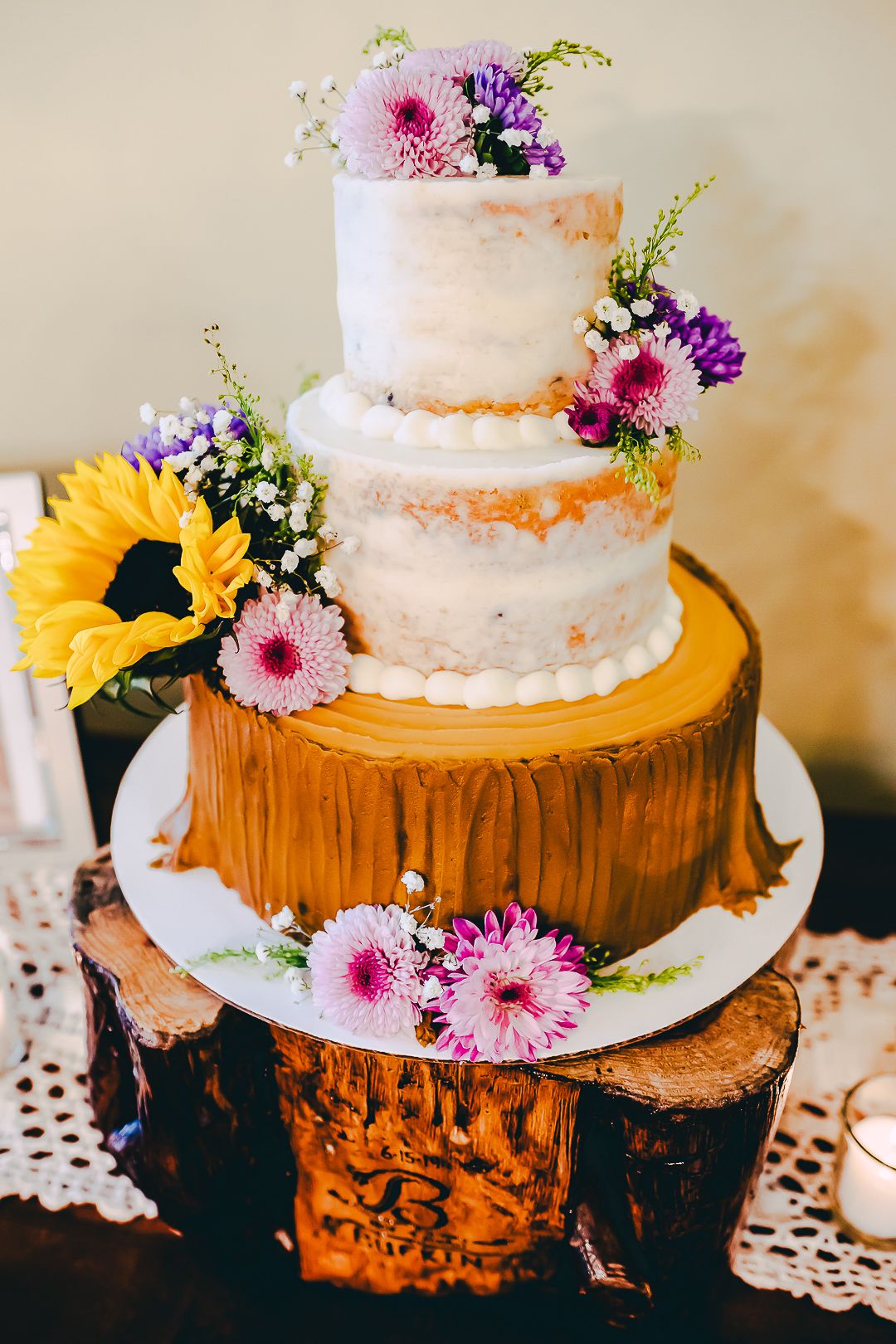 Rustic wedding cake decorated with fresh flowers sits on wooden log slice cake stand with lace doily.