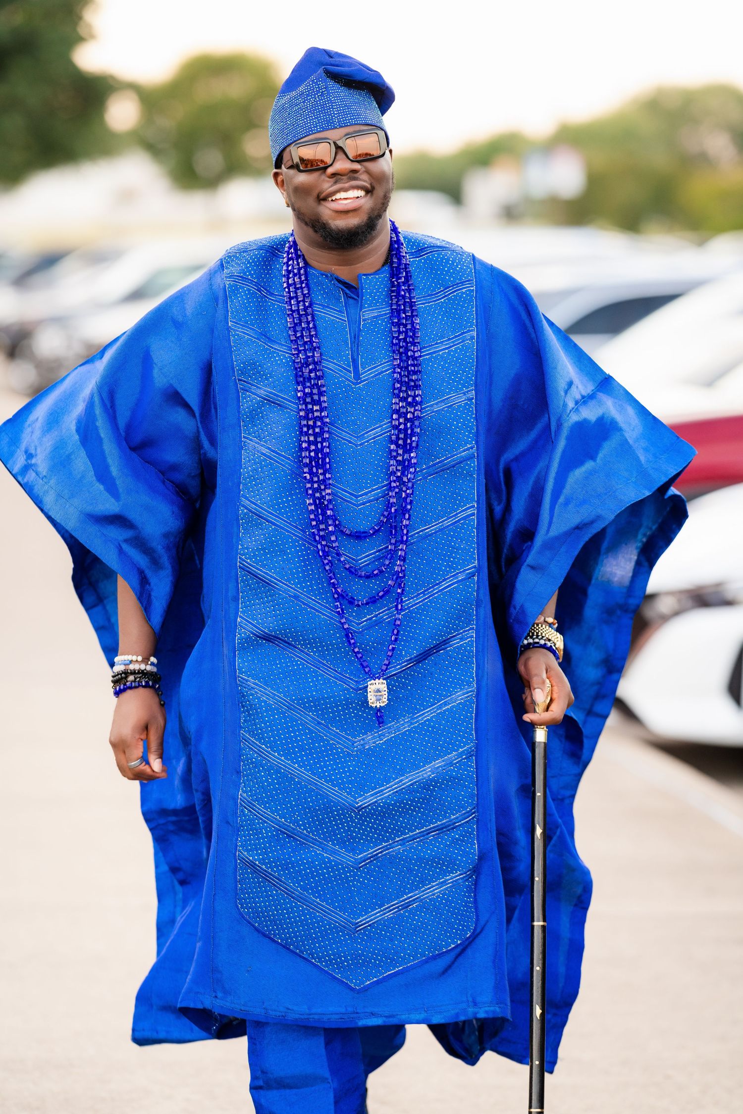 Person wearing traditional bright blue agbada with beaded necklace outdoors.