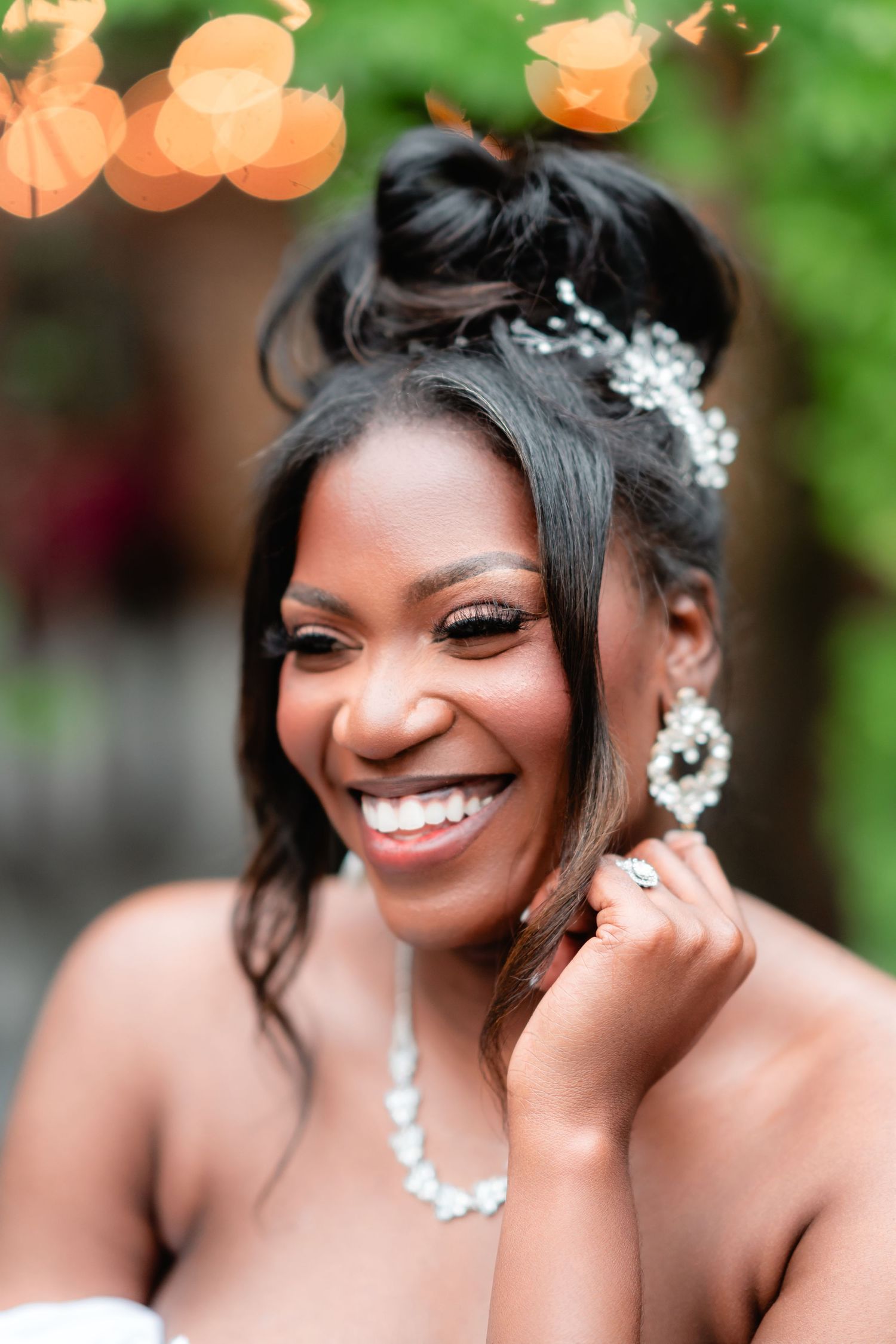 A radiant bride wearing a floral hair accessory and pearl jewelry smiles joyfully against a green outdoor backdrop.
