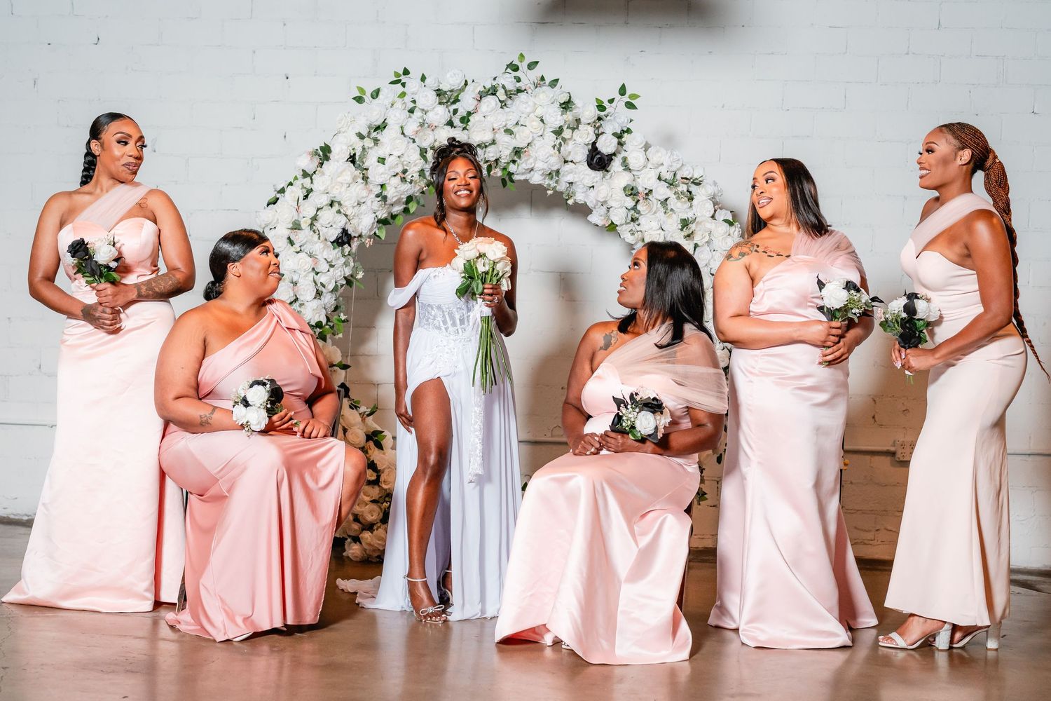 Bridesmaids in blush pink dresses pose in a row against a white floral backdrop at an elegant wedding.