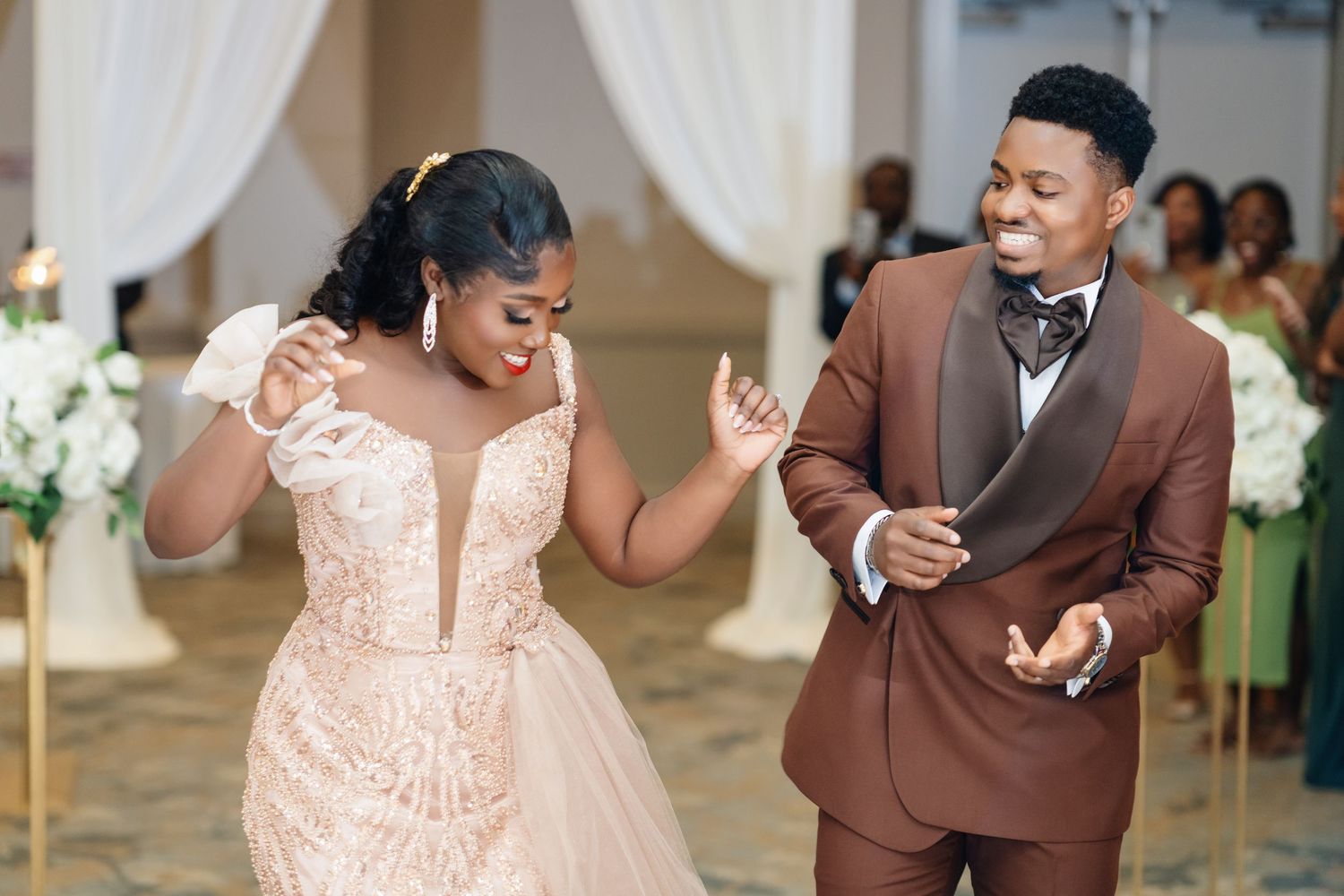 A couple in brown and blush pink attire perform a choreographed dance routine at an elegant indoor venue.