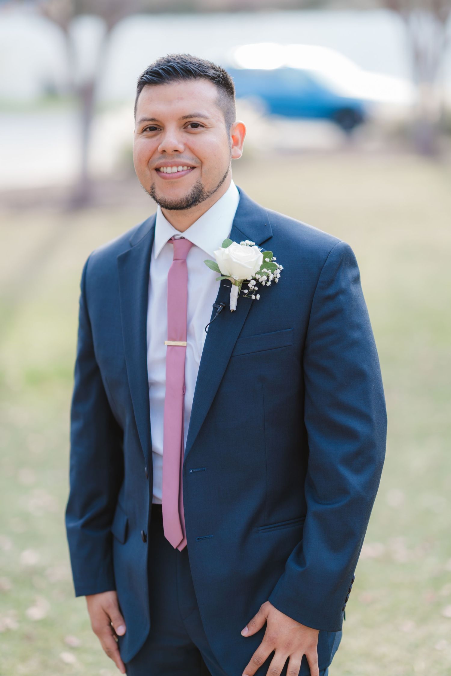 Groom wearing navy suit with pink tie and white boutonniere poses in outdoor setting.