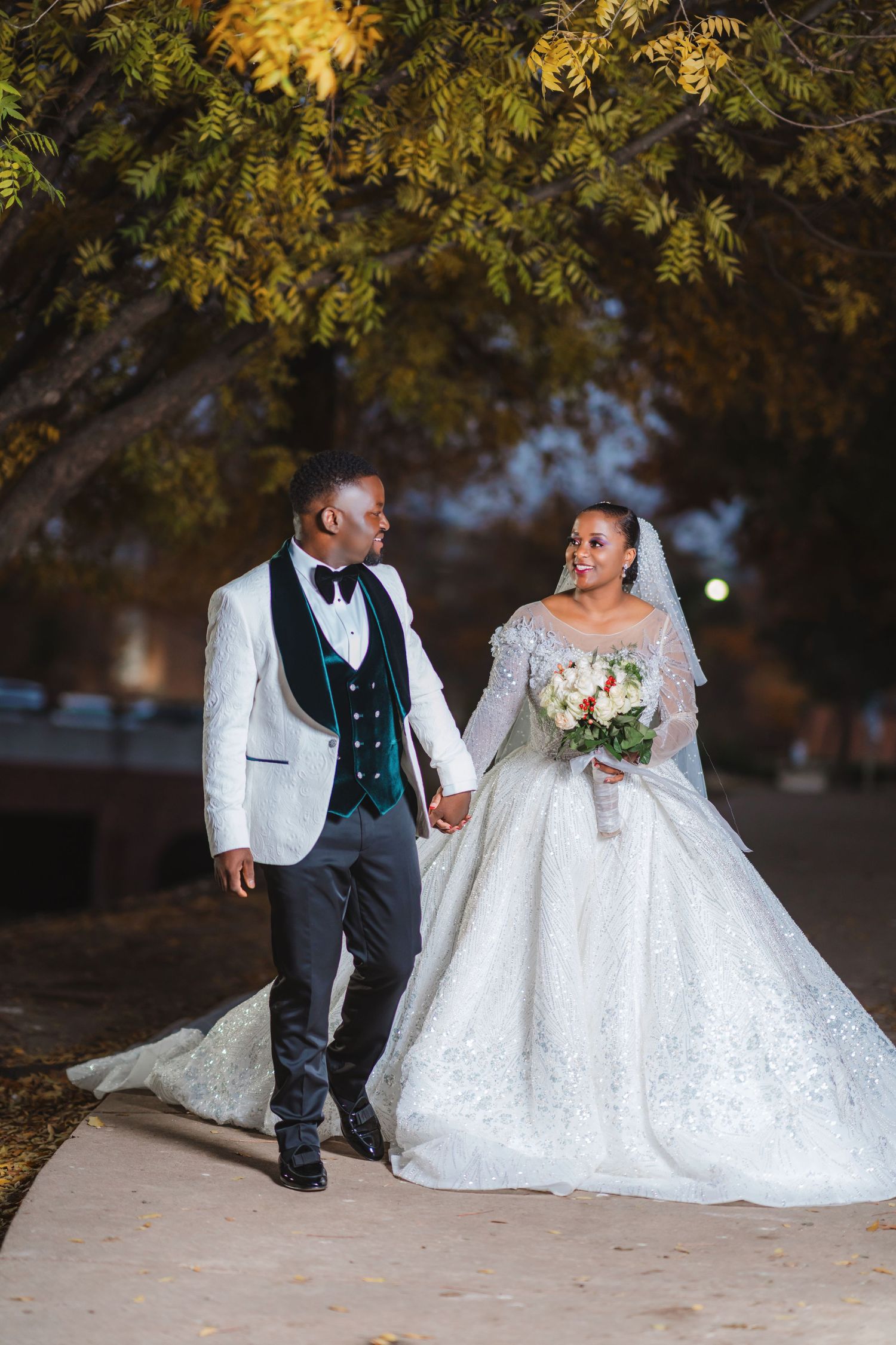 A romantic wedding portrait of a couple walking under autumn trees in formal attire.