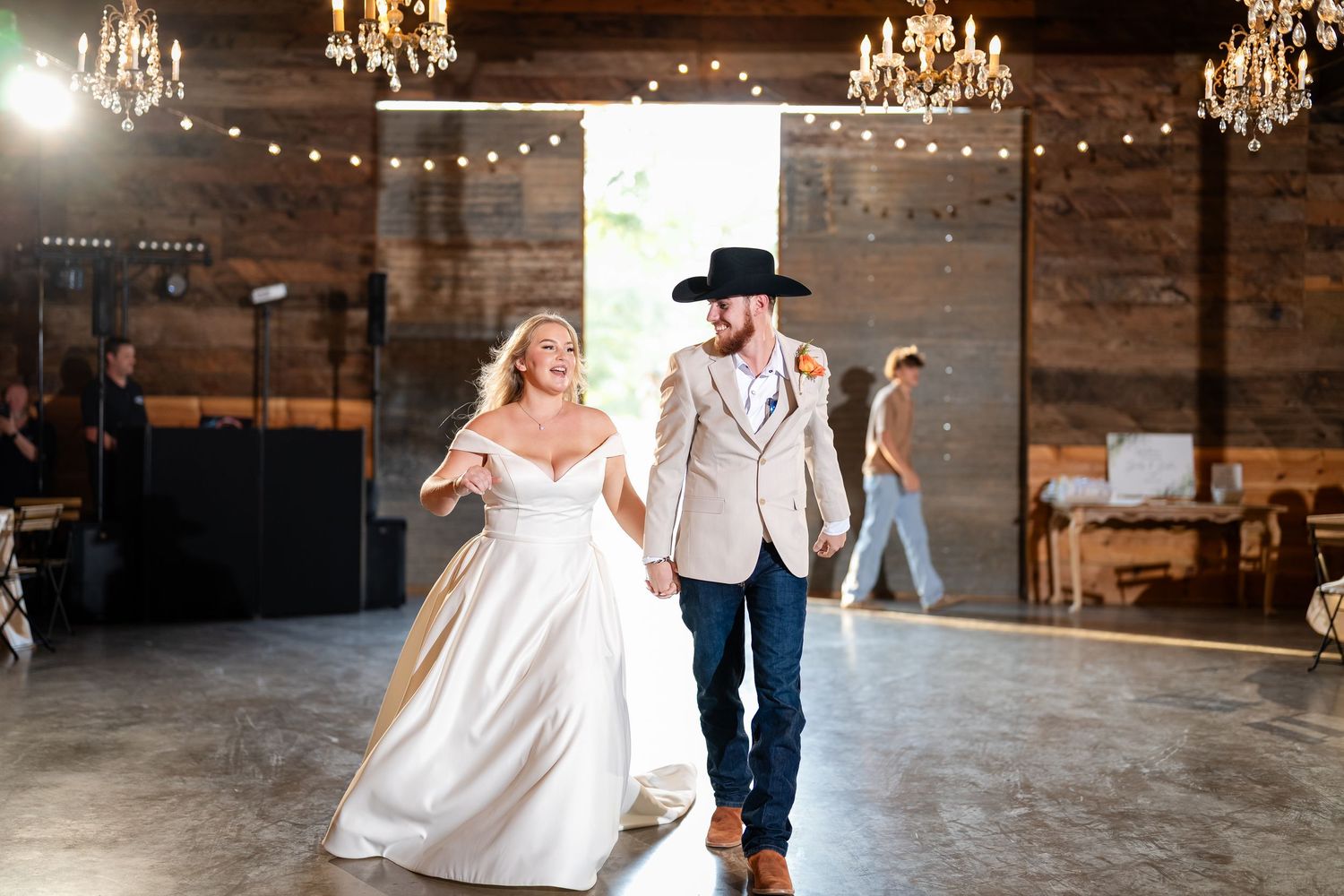 A couple dances in an elegant barn venue decorated with chandeliers and string lights.