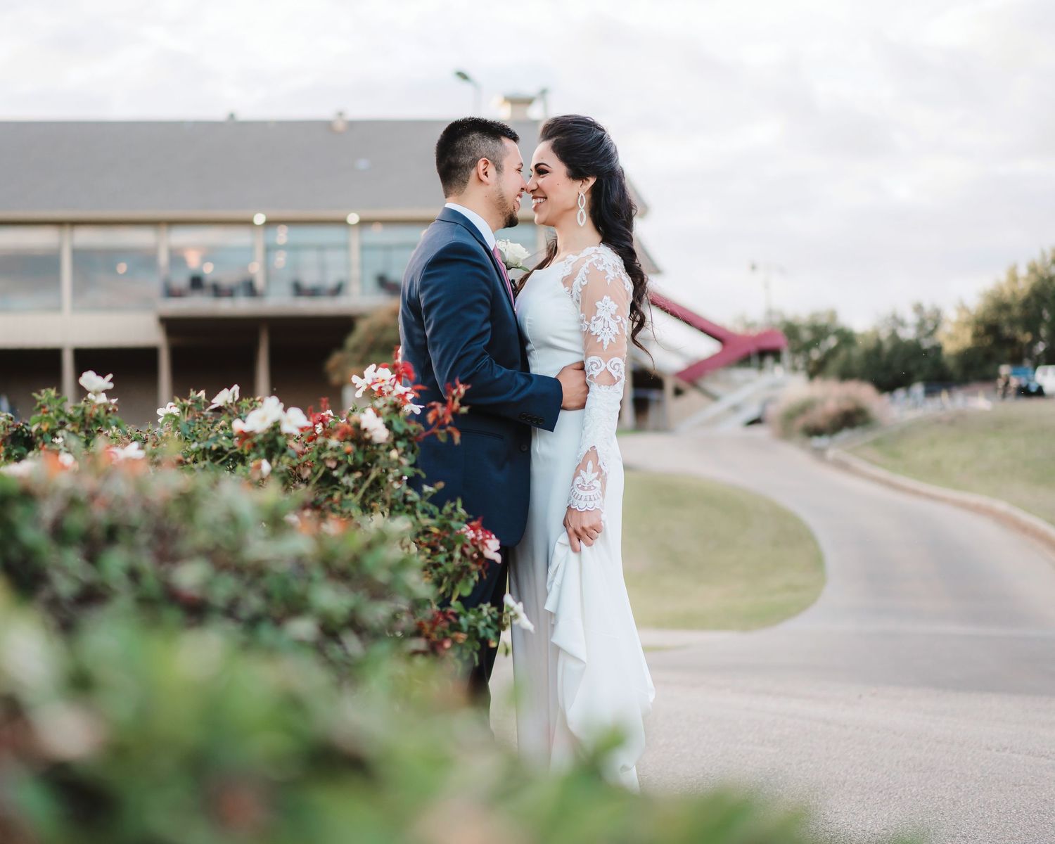 Romantic couple in wedding attire embrace while walking along a scenic outdoor path at sunset.