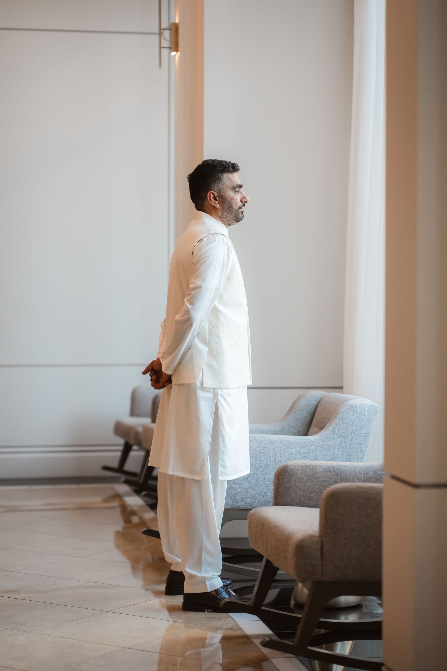 A person in white formal attire stands in a minimalist room with gray furniture.