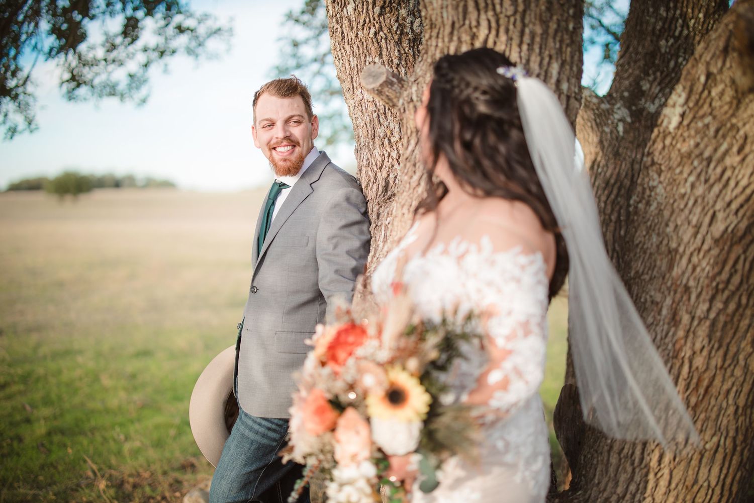 Wedding couple shares intimate moment under large tree with coral and peach floral bouquet.