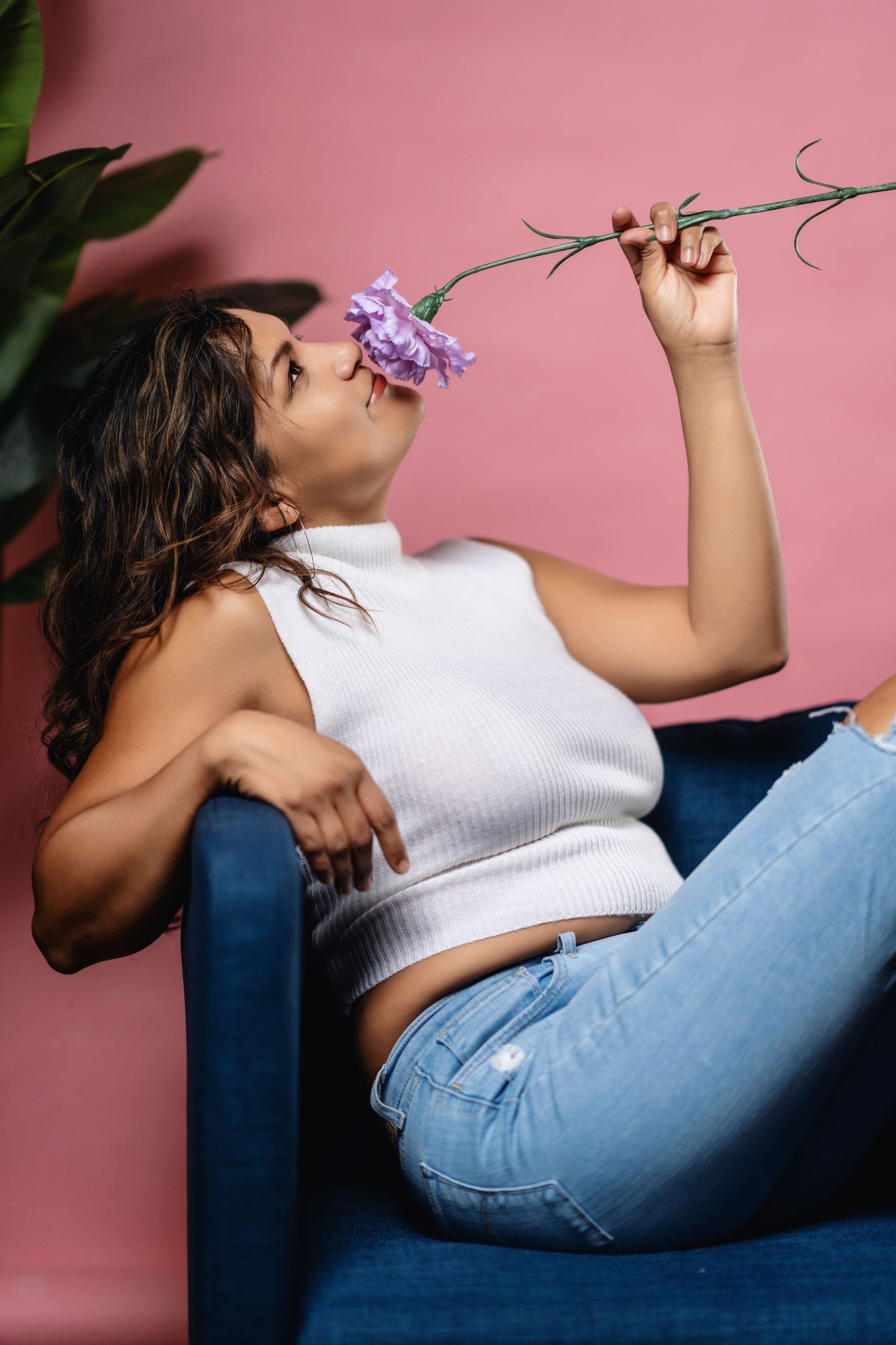 Someone in a white crop top and jeans lounges on a blue chair smelling a purple flower against a pink wall.