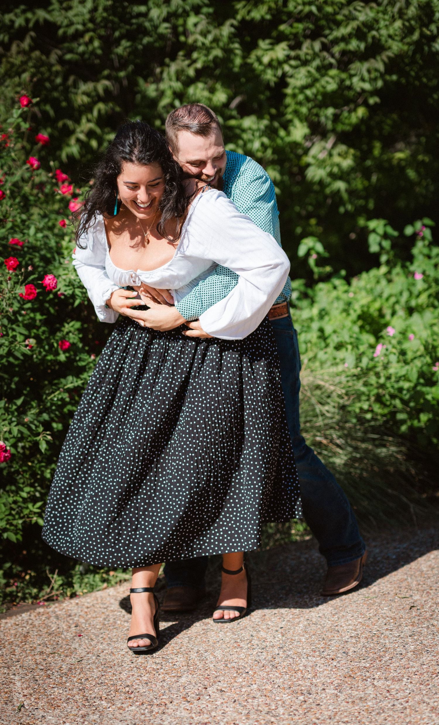 A romantic couple embraces on a garden path surrounded by red roses and greenery in a candid outdoor photoshoot.