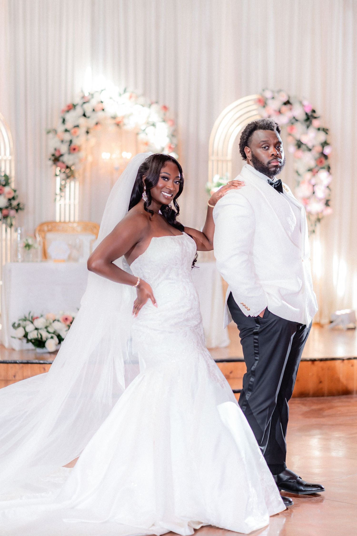 A couple in wedding attire dances and twirls in an elegant ballroom decorated with floral arrangements.