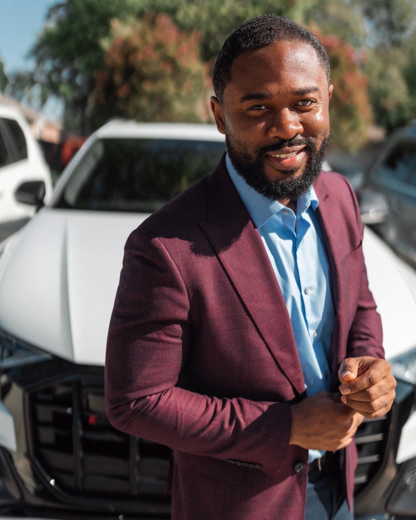 A person in a burgundy suit jacket and light blue shirt smiles warmly while standing outdoors.