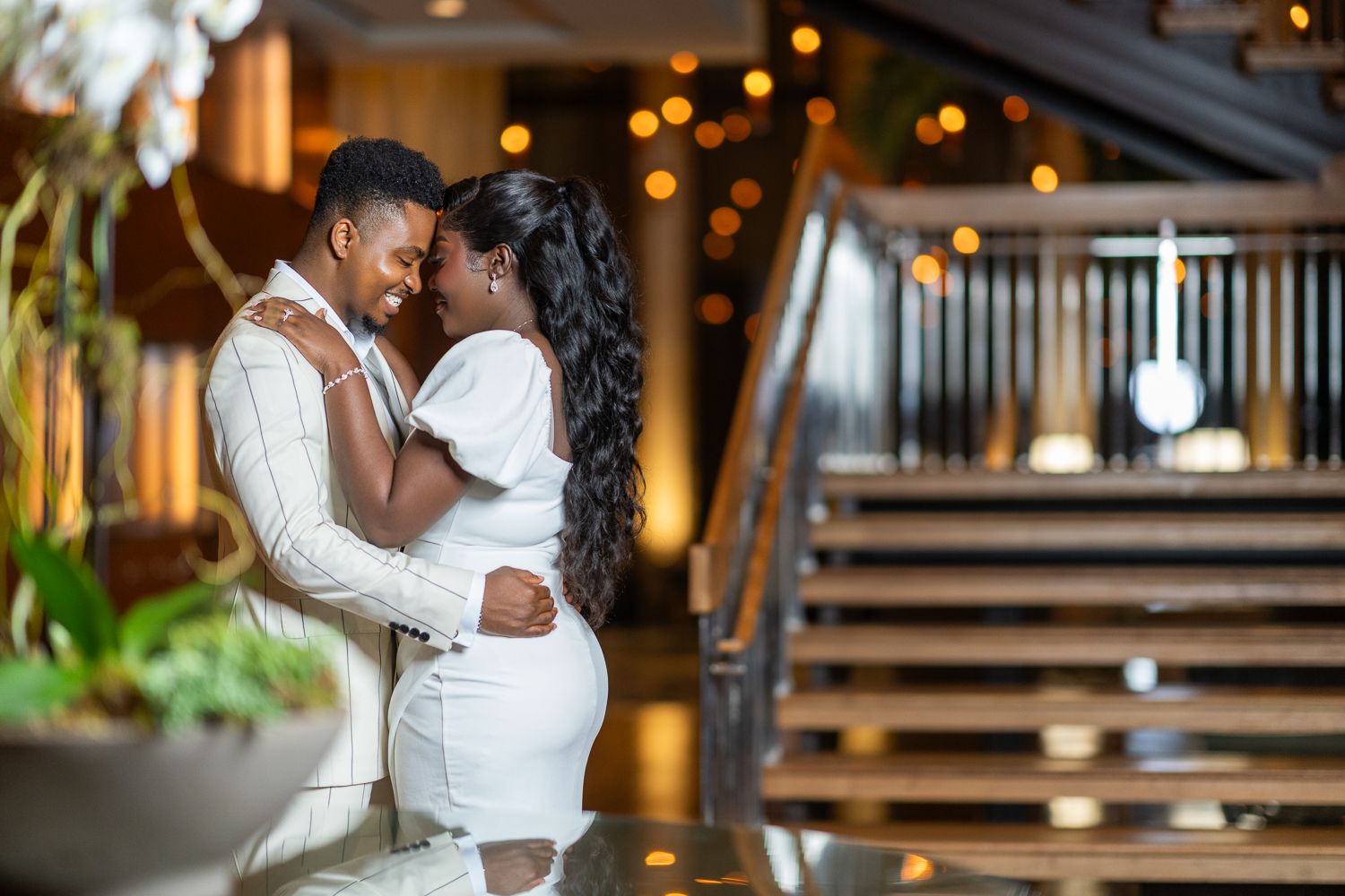 A couple embraces in front of warm glowing lights and wooden stairs during an evening photoshoot.
