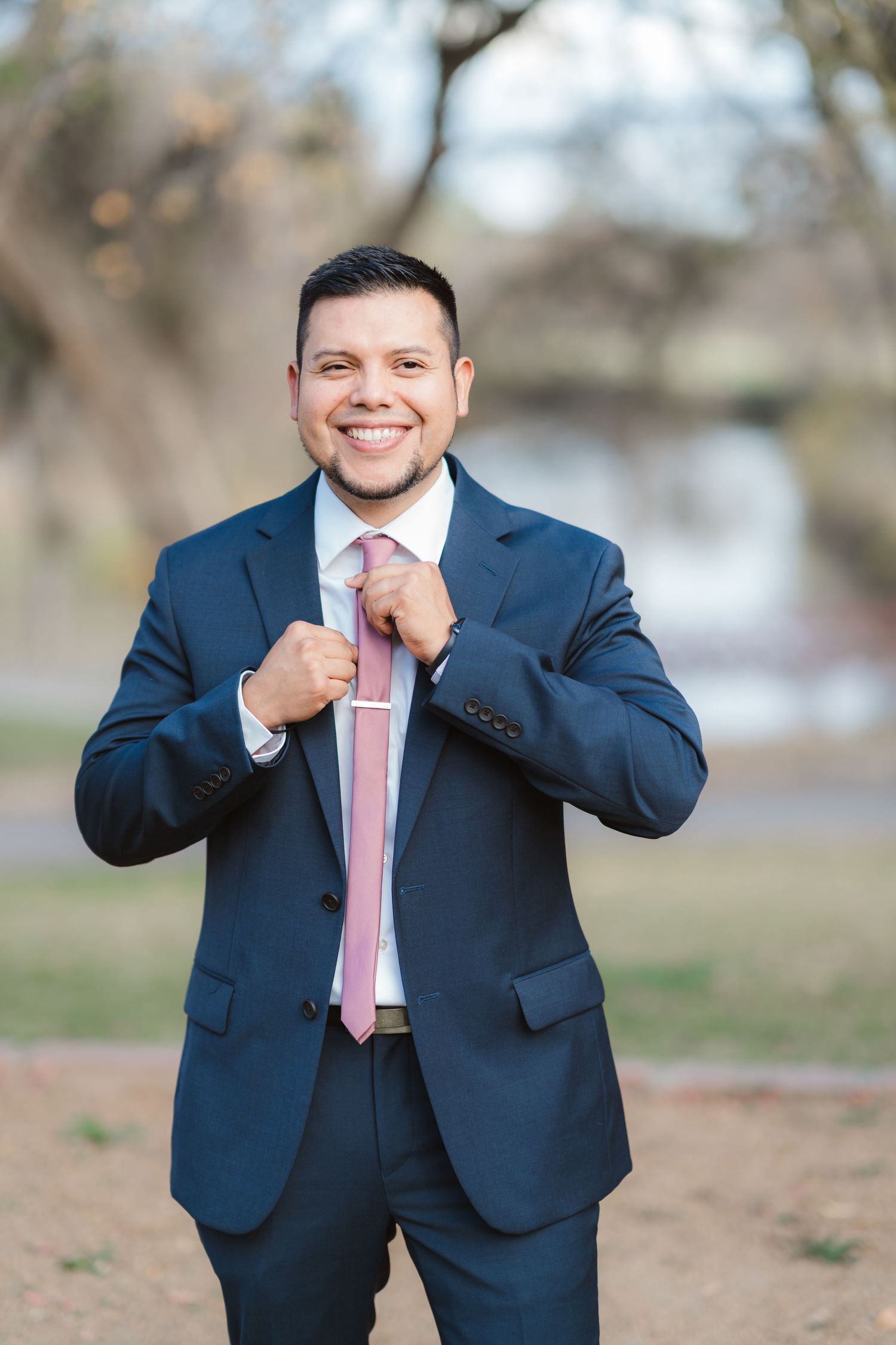 A groom in a navy suit and pink tie poses playfully in an outdoor setting.
