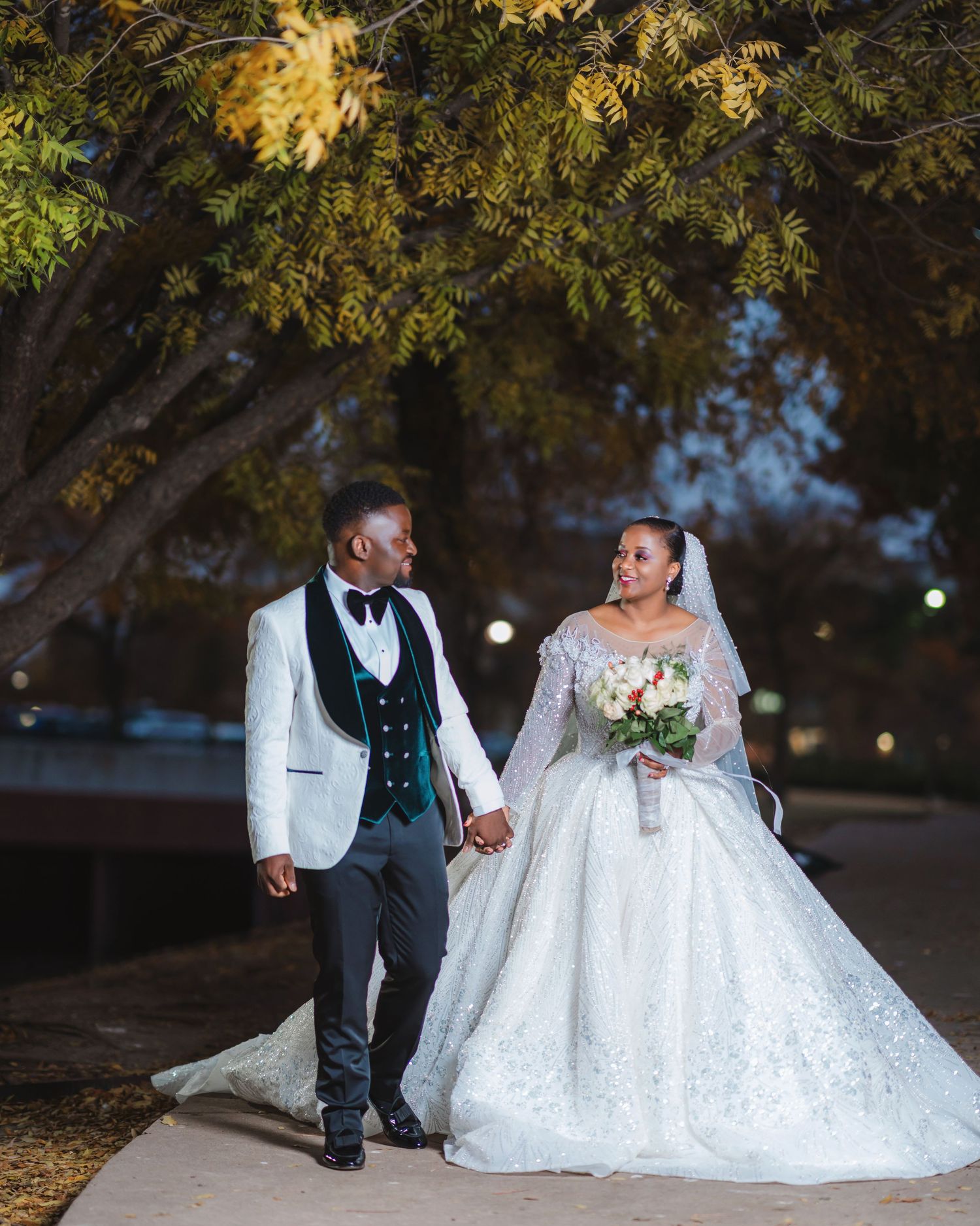 A wedding couple walks together under trees at dusk, with the bride wearing a flowing white gown and groom in white jacket.