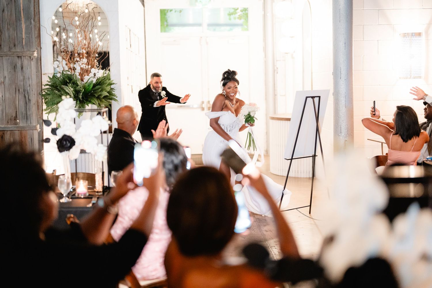 Wedding guests raise their hands in celebration during a joyful reception moment at an indoor venue.