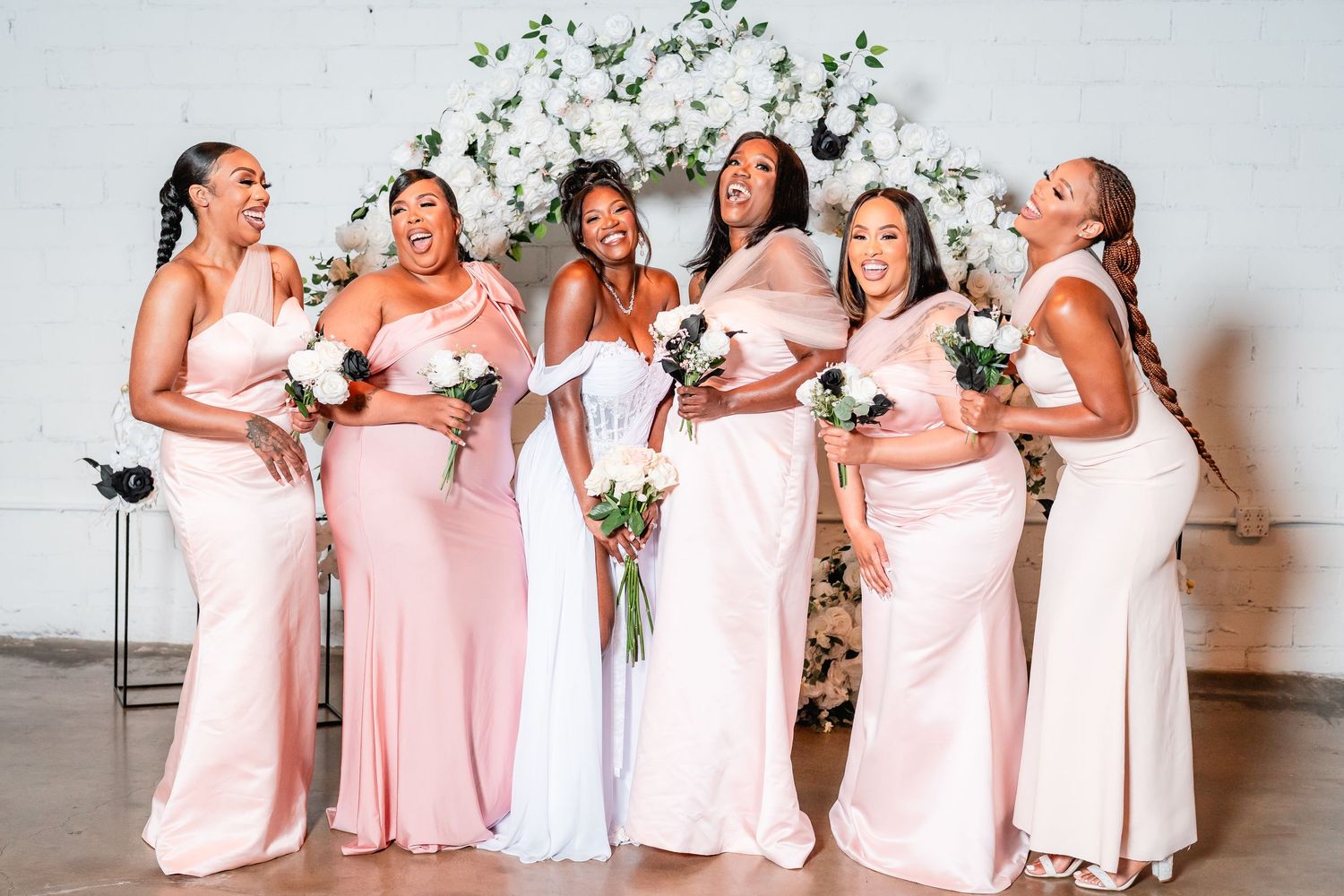 Bridesmaids in light pink dresses laugh and celebrate together against white backdrop.