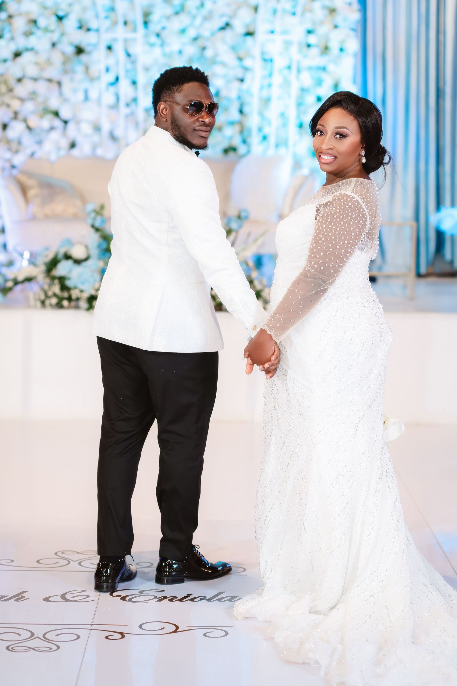 Wedding couple in white dress and black suit pose together on decorated white floor with blue backdrop.