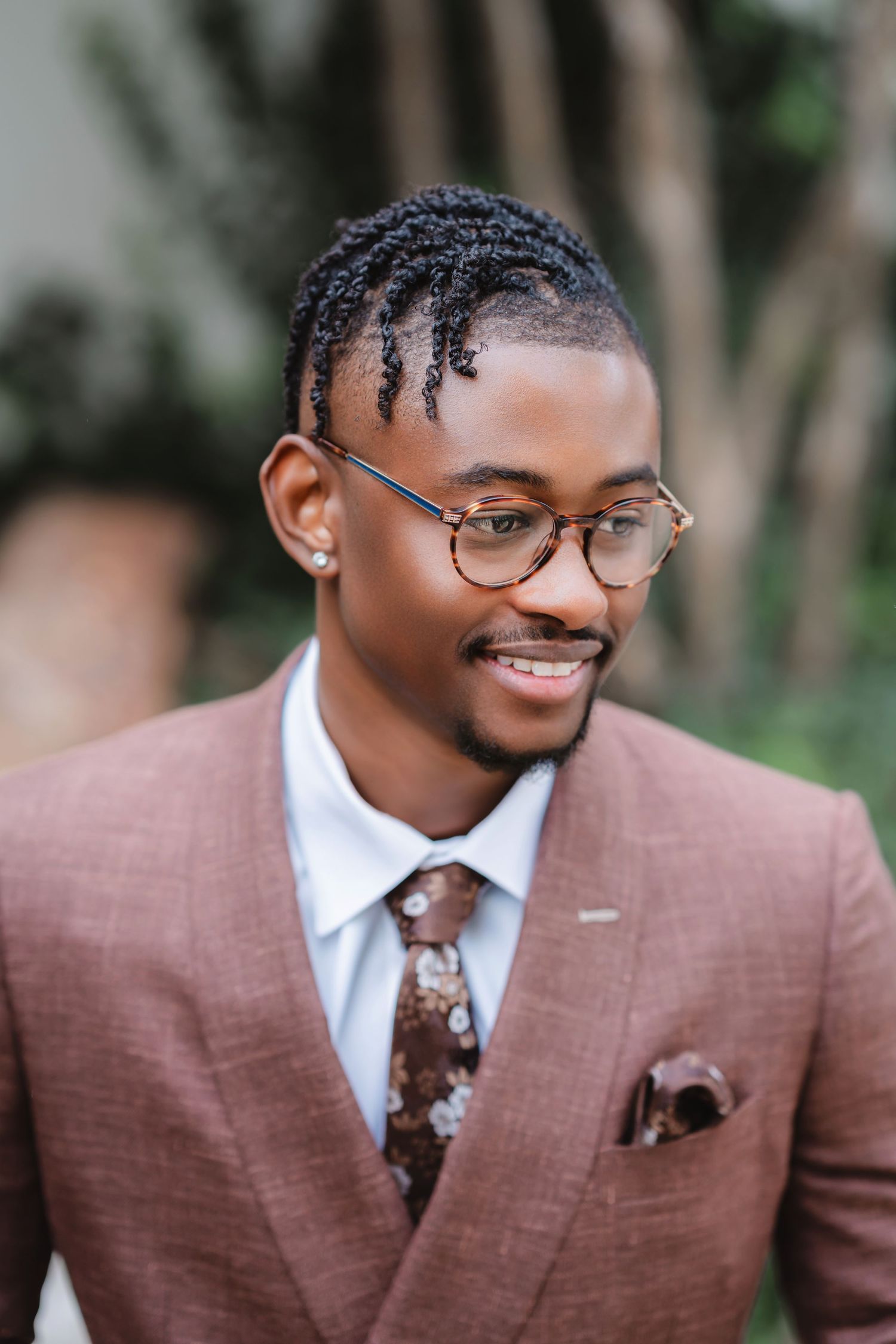 Groom in a stylish brown suit and glasses smiles warmly during a wedding photoshoot outdoors.