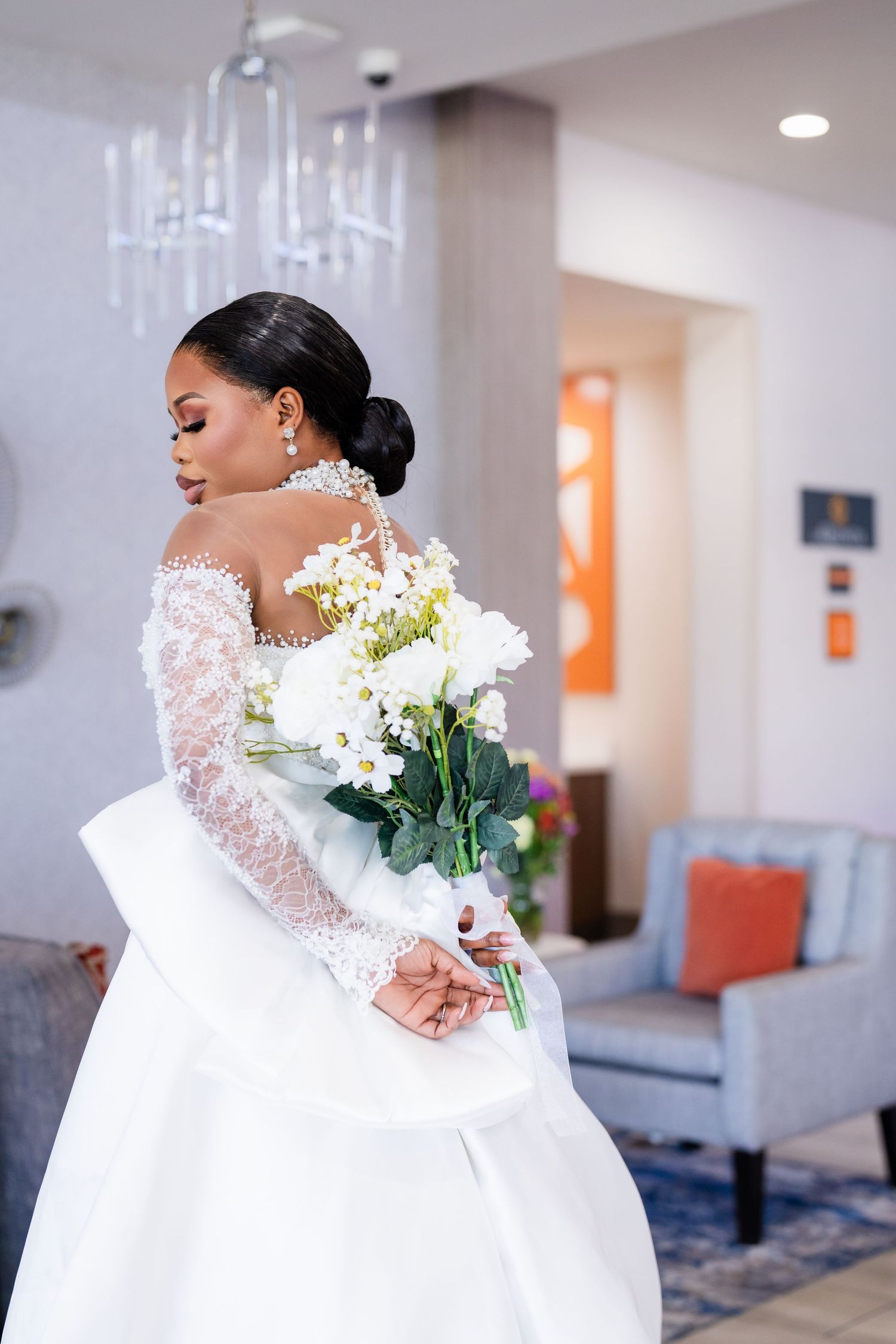 A bride in an elegant lace-sleeved wedding gown holds a white floral bouquet in an upscale venue with chandeliers.