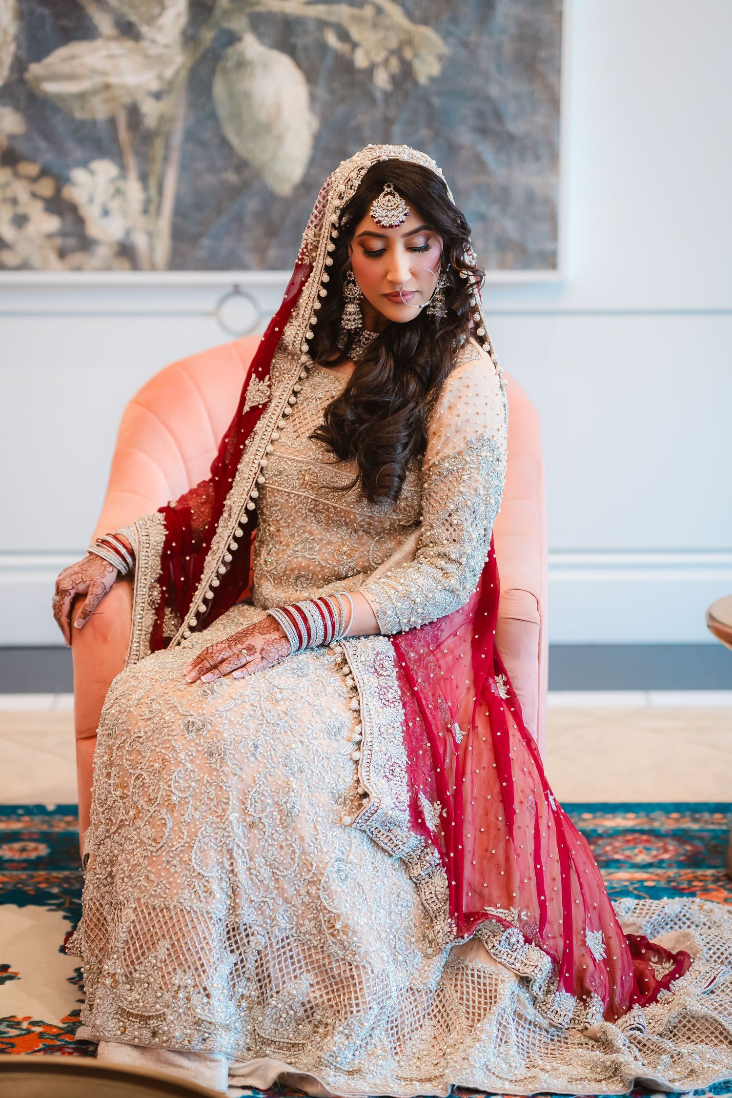 Traditional South Asian bride in an ornate beige and silver gown with red dupatta sitting on an orange couch.