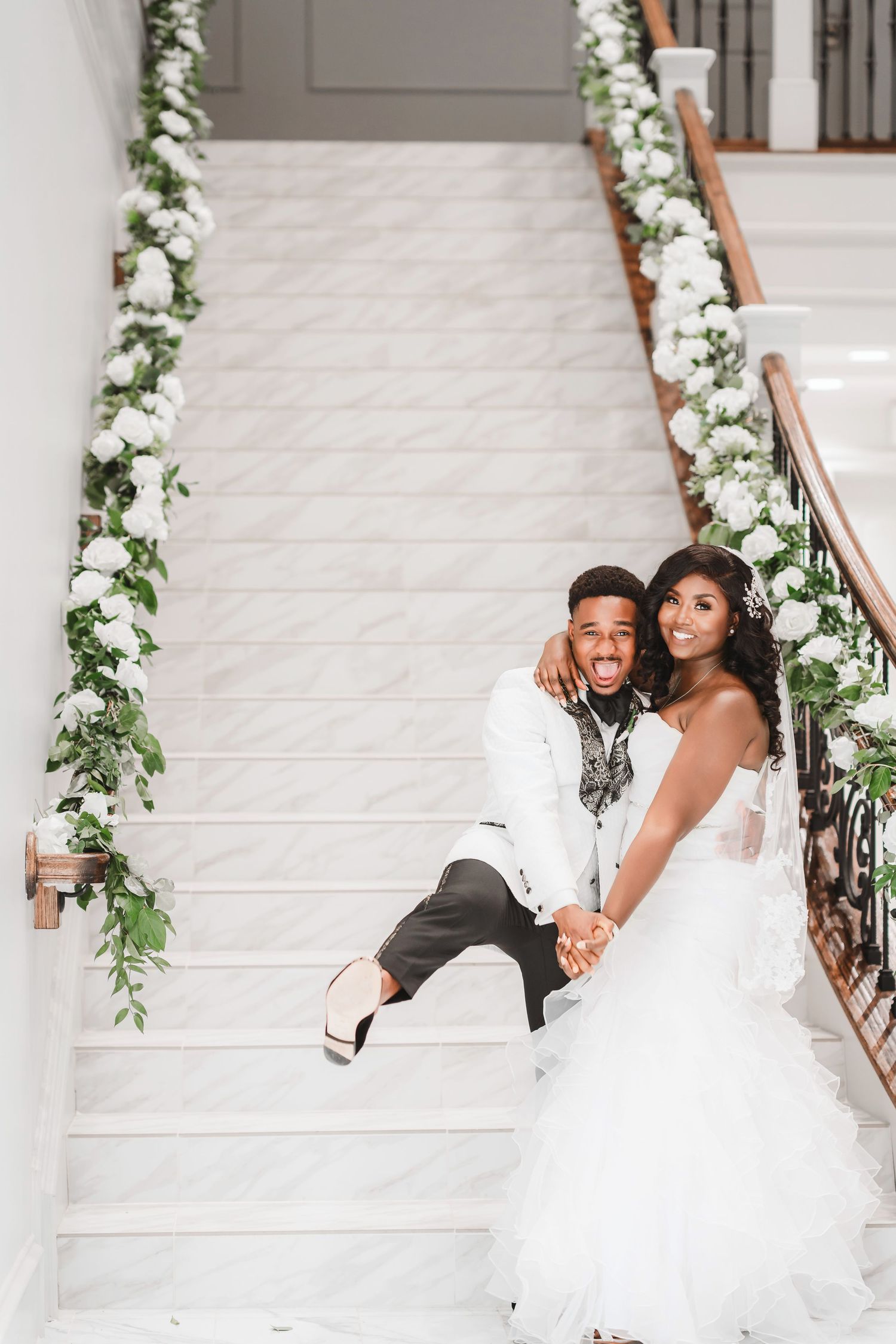 Playful wedding couple poses on white staircase with floral garland decorations.