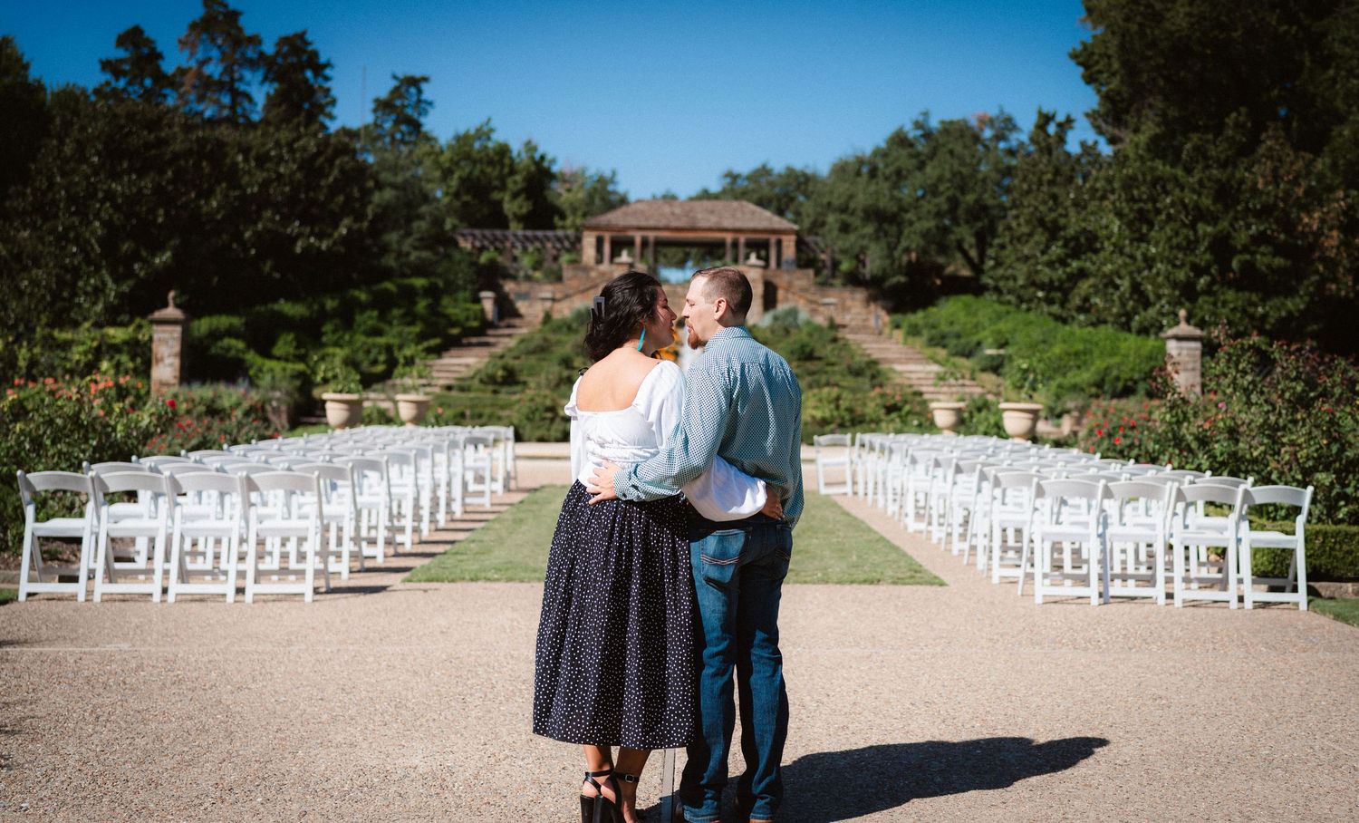 White chairs line a garden pathway under blue skies for an outdoor wedding ceremony.
