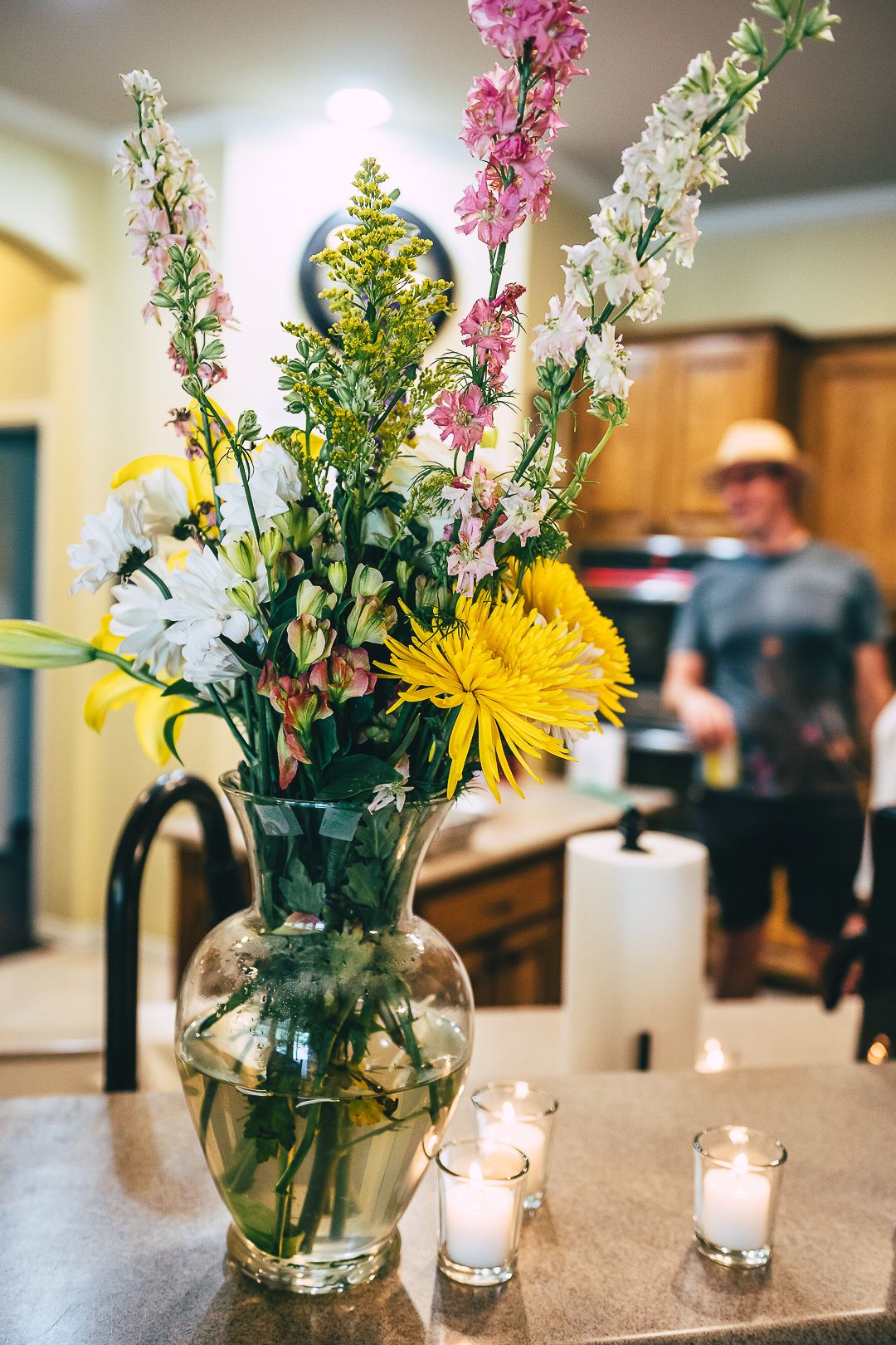 Floral centerpieces with pink and yellow flowers in glass vases alongside lit candles on a counter.
