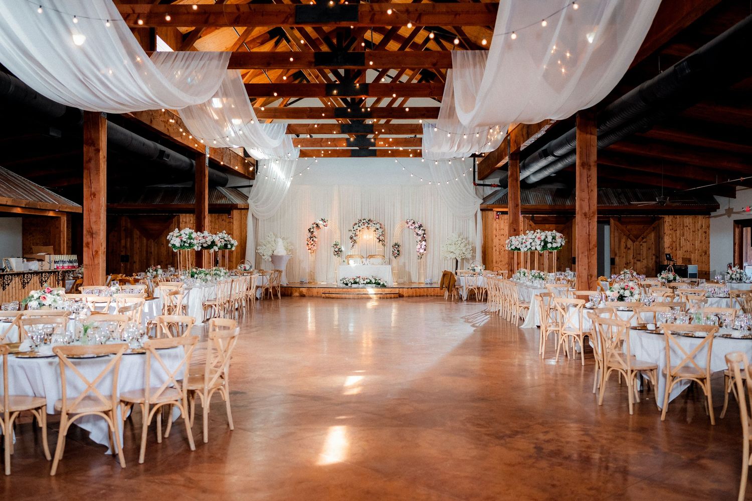 Elegant rustic barn wedding reception venue decorated with white drapes, chandeliers and round dining tables.