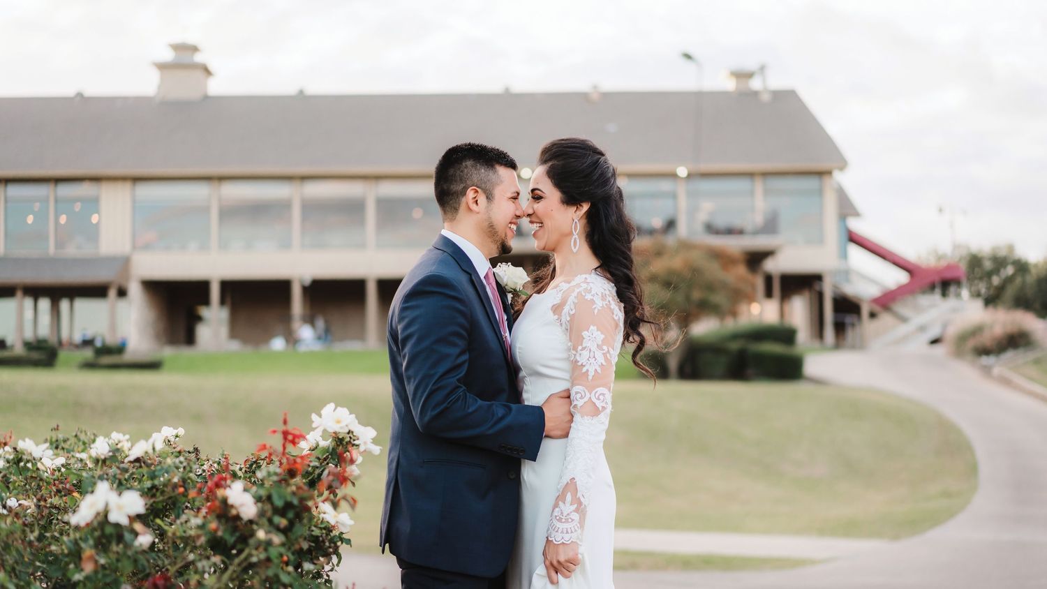 Romantic outdoor wedding couple embraces in front of rustic barn venue with floral arrangements.