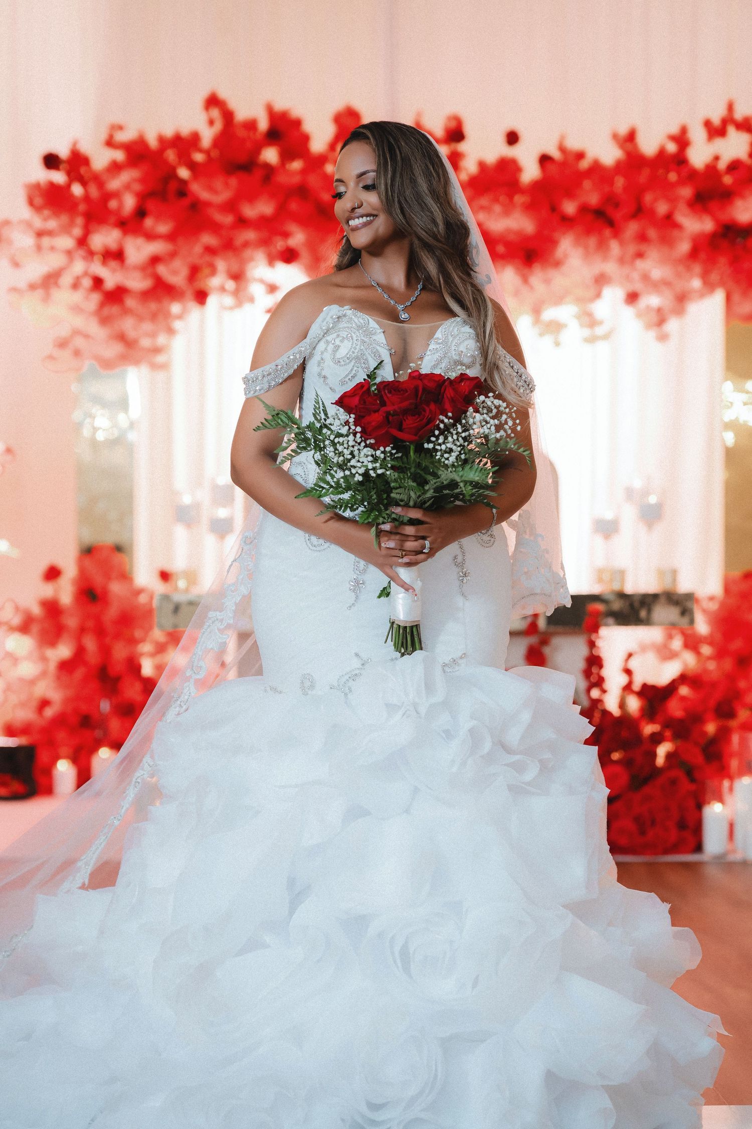 A bride wearing a ruffled white wedding dress smiles while holding a red rose bouquet against a red floral backdrop.