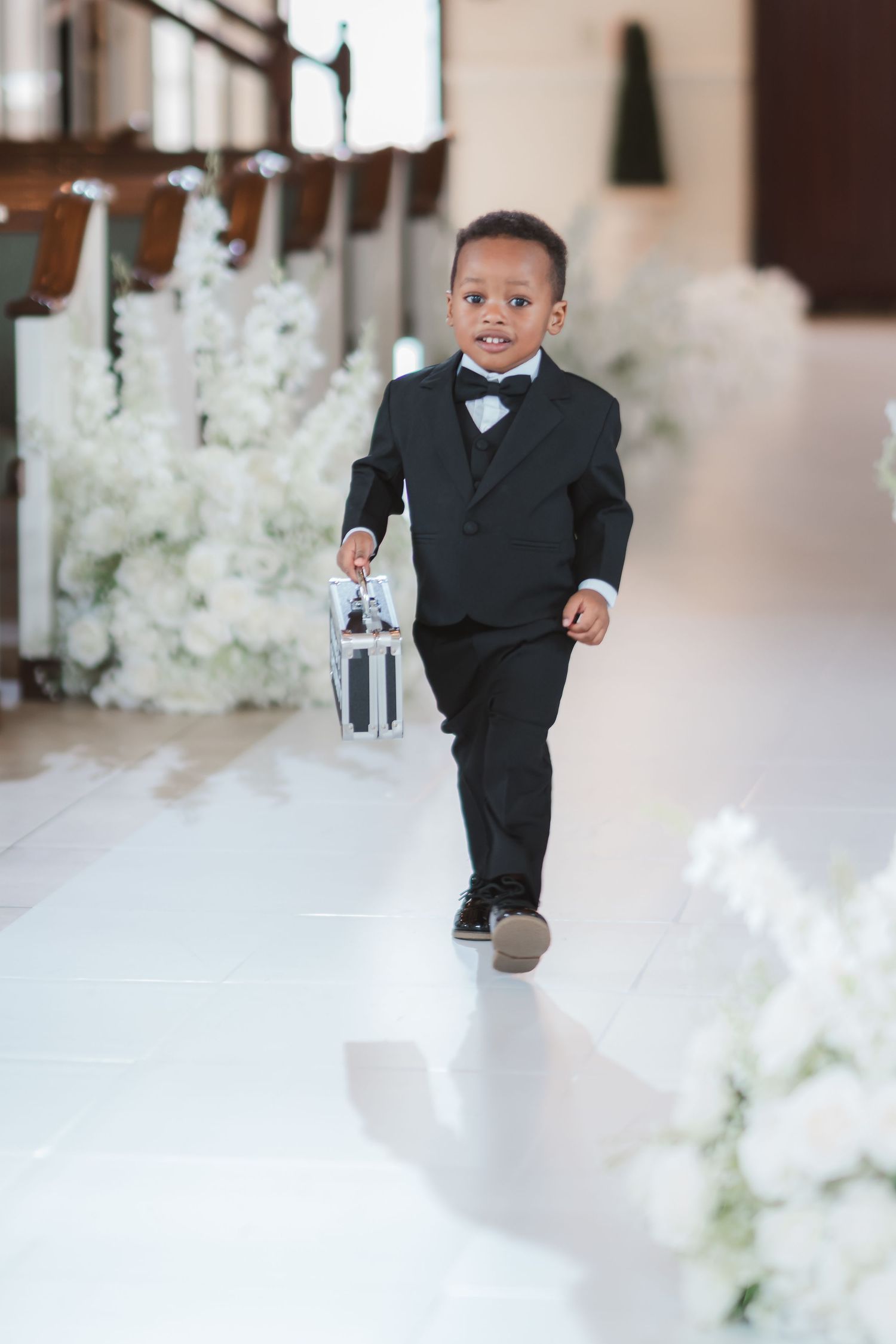 Young ring bearer in a black suit walks down the aisle at an elegant wedding ceremony with white floral decorations.