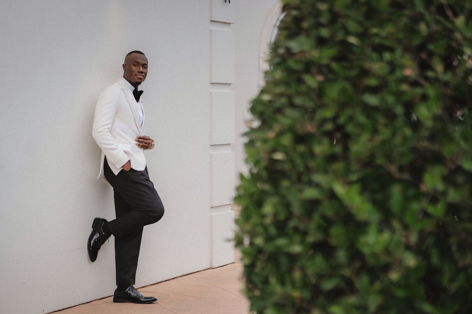 A small child in formal attire poses against a white wall during a wedding event.