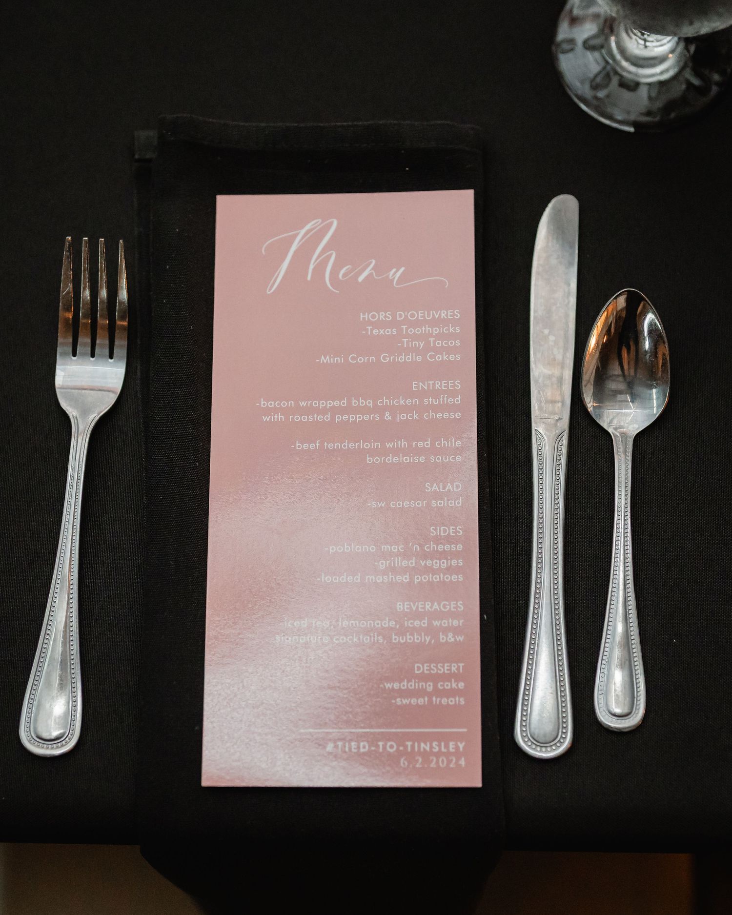 Wedding menu cards in blush pink displayed with elegant silver flatware on a black tablecloth.