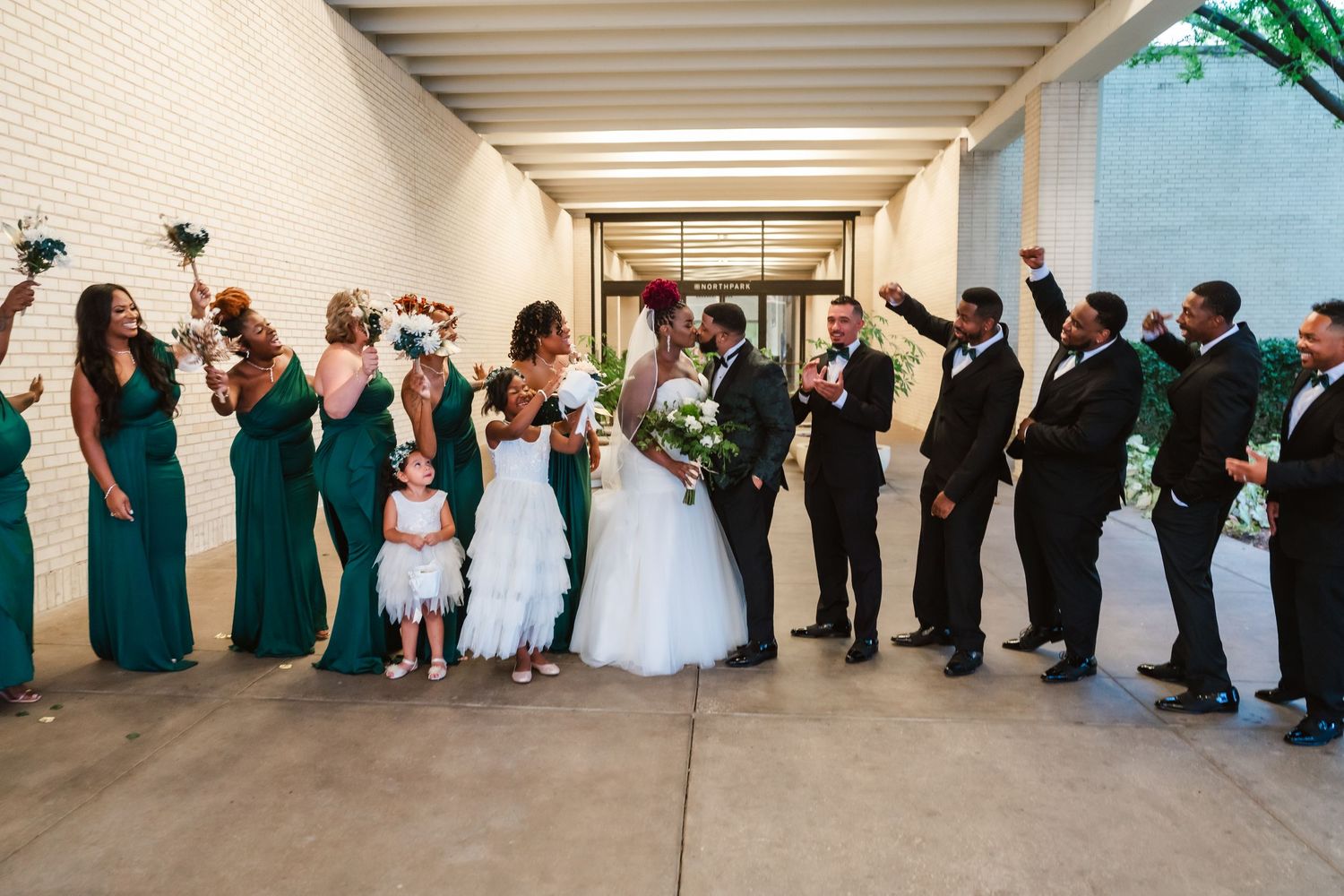 Large wedding party lined up for a formal group photo in front of a modern architectural backdrop.