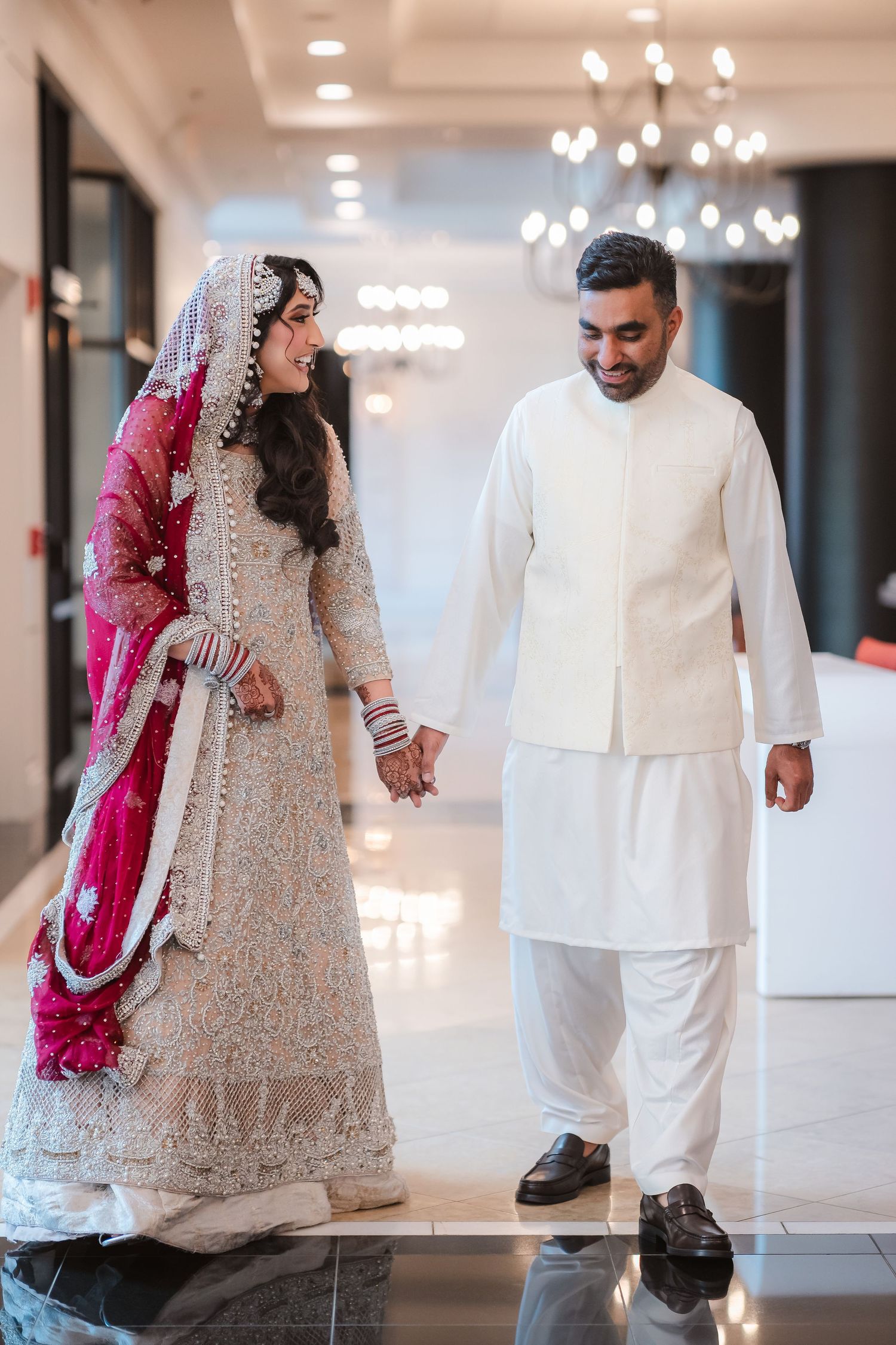 Couple walking down an elegant hallway with chandeliers wearing traditional South Asian wedding attire.
