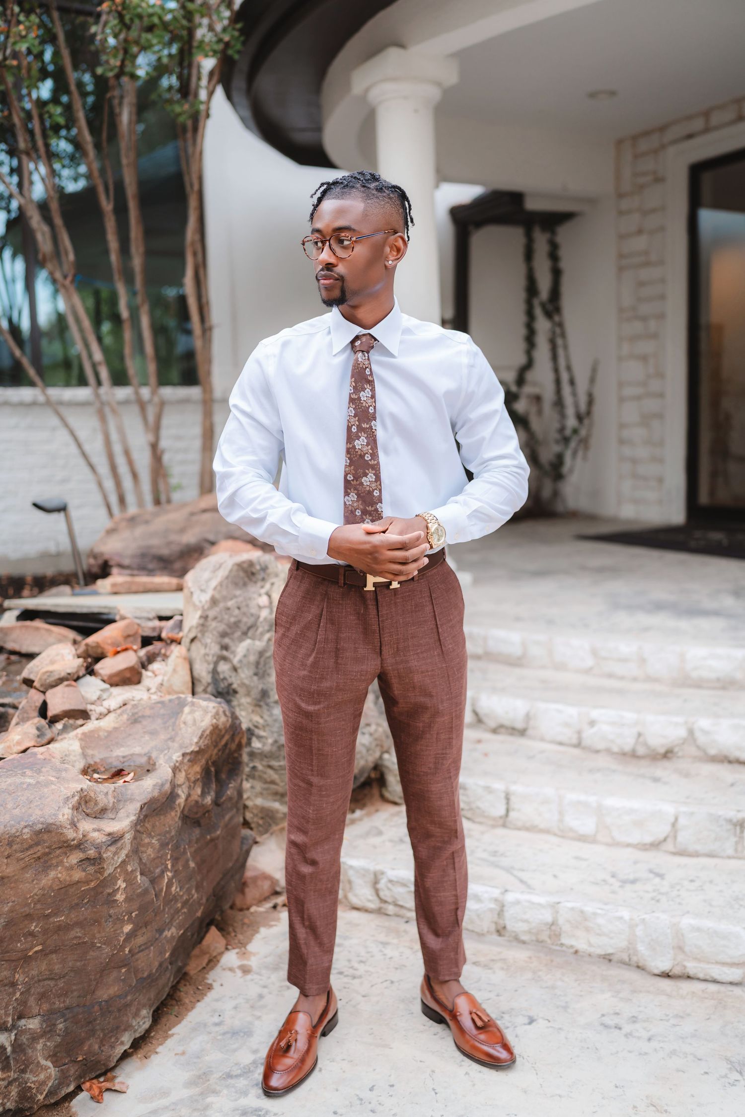 Person in formal attire poses against white building exterior with brown pants and tie.
