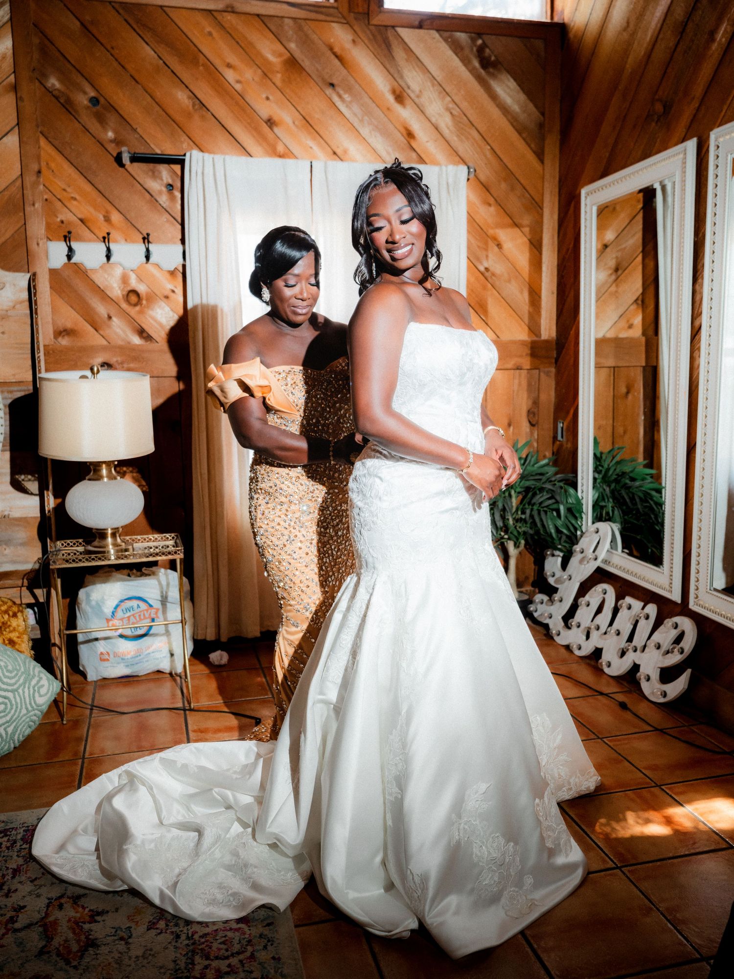 Two people pose in a rustic wood-paneled room, one wearing a white wedding gown and another in a gold sequined dress.
