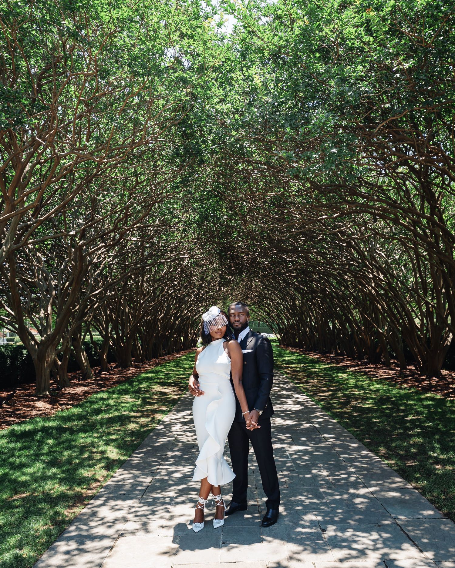 A couple walks through a serene garden pathway lined with dense green shrubs on a sunny day.