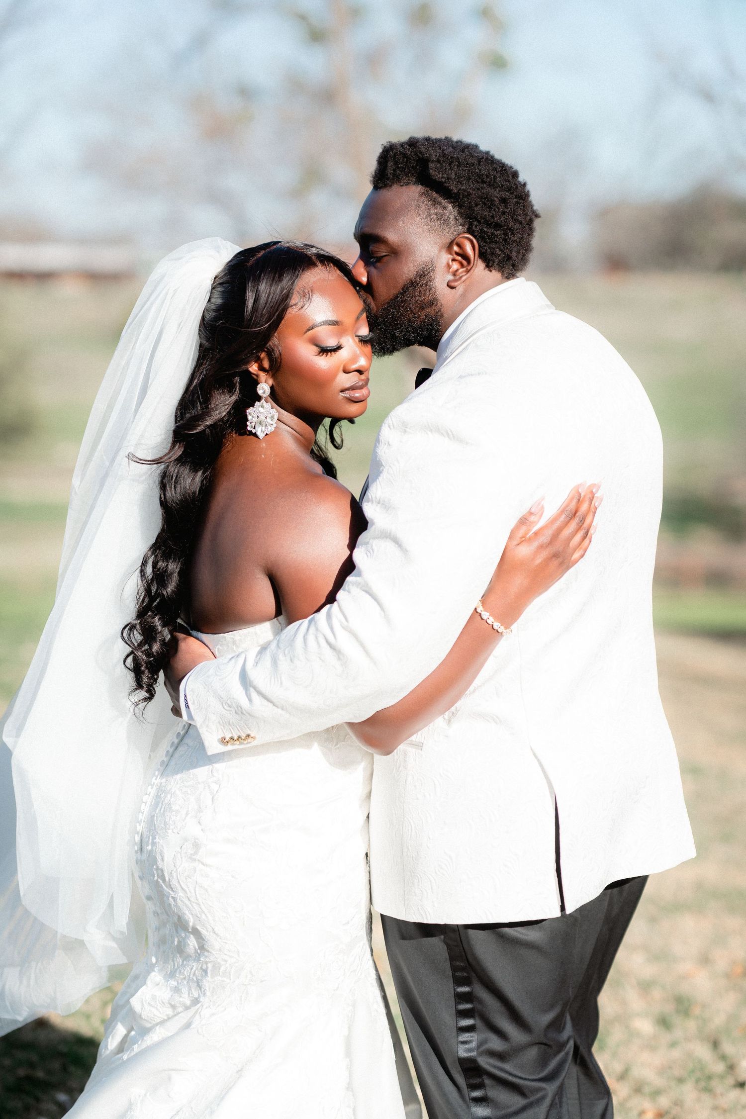 Intimate moment between newlyweds in white attire sharing an embrace outdoors during their wedding photoshoot.