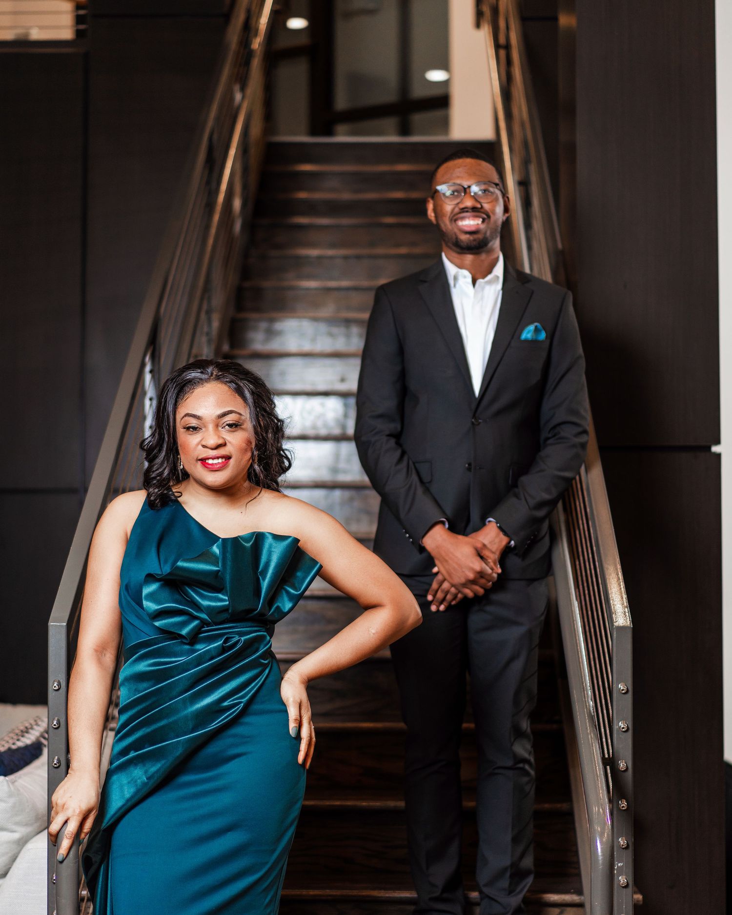Elegant couple in formal attire poses on dark staircase with teal velvet dress and black suit.