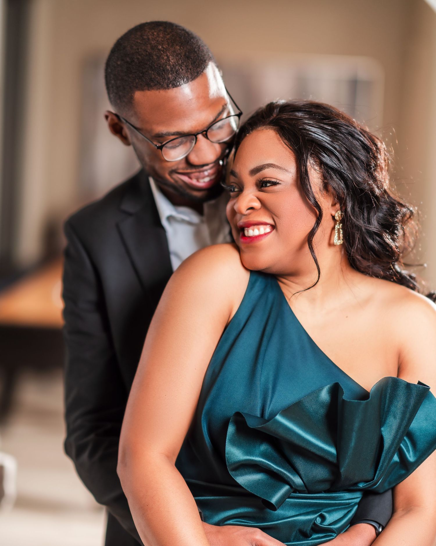 A couple in formal attire share a tender moment with the person in a teal dress leaning back while laughing.