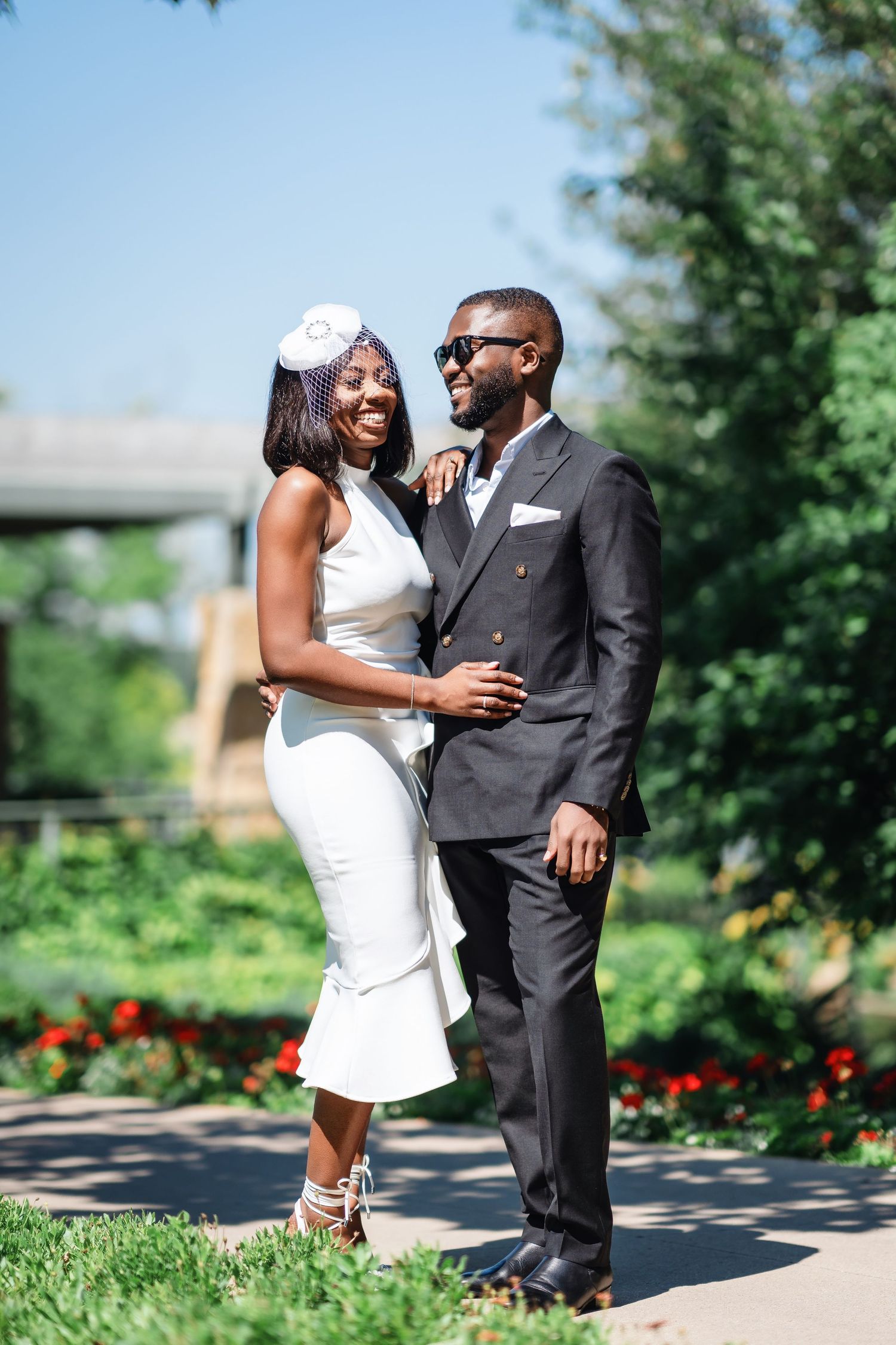 A romantic couple poses outdoors in formal attire against a garden backdrop with blooming flowers.