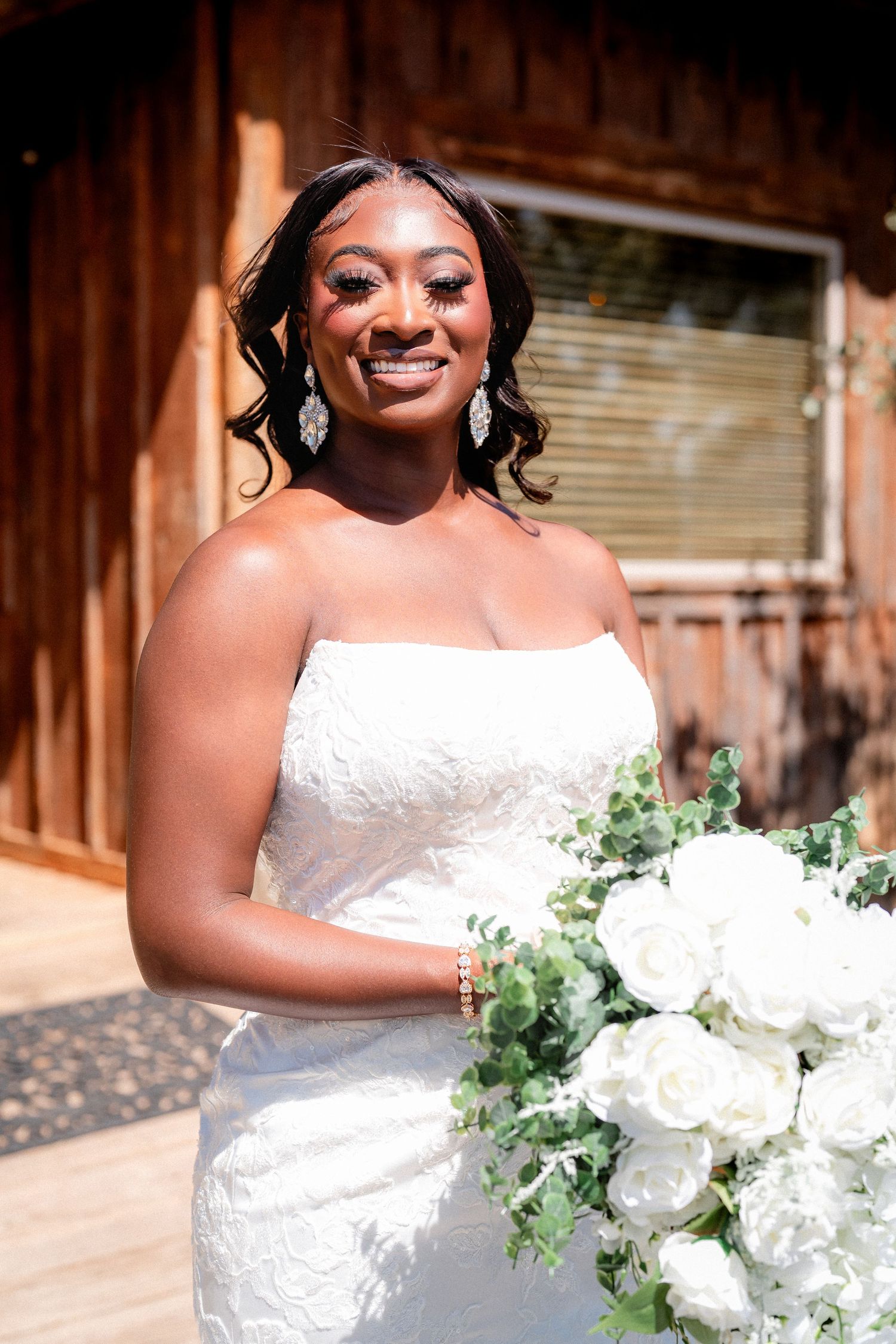 A bride in a white lace gown holds a bouquet of white flowers against a rustic wooden backdrop.