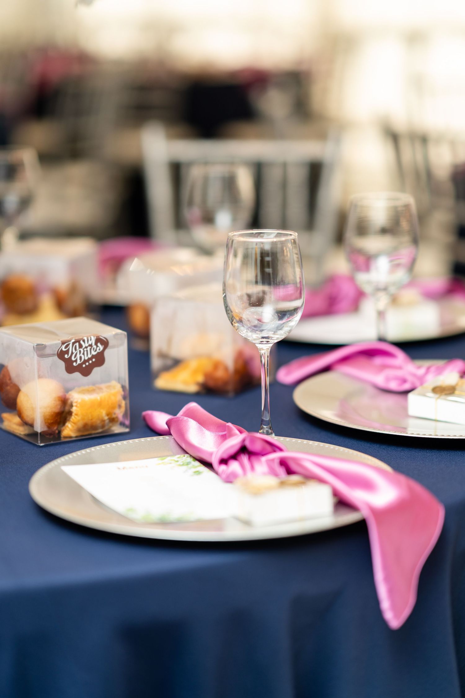 Table settings at a formal event with navy blue tablecloths, pink napkins and wine glasses arranged in a row.